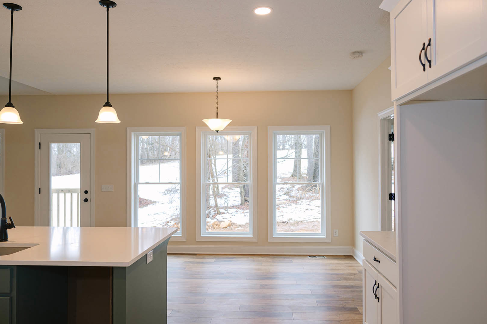 Spacious kitchen featuring a large white island with black and grey base, wooden flooring, white walls, cabinetry, and expansive windows overlooking a snowy forest.