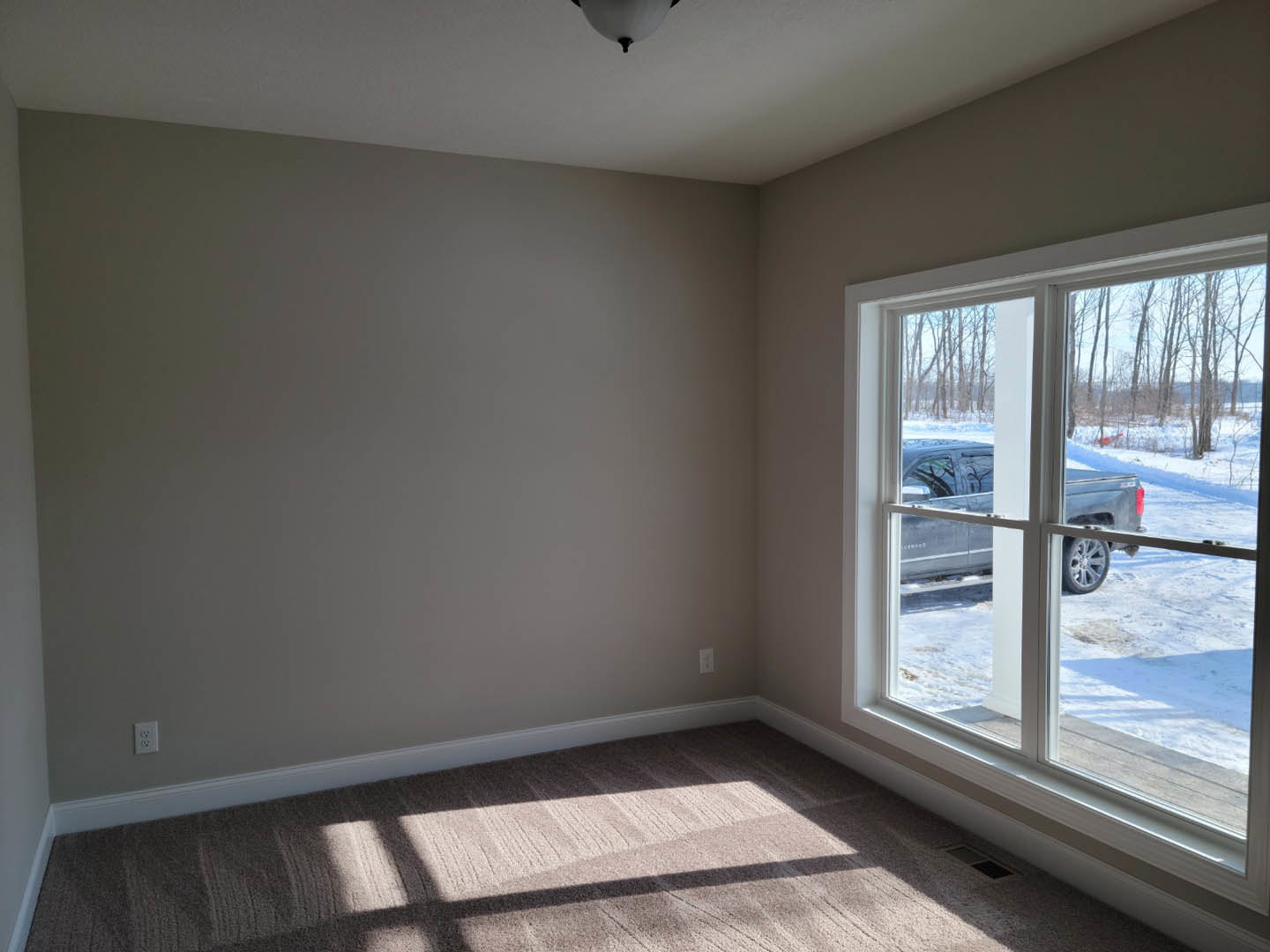 Carpeted room with white ceiling and walls, large window overlooking snowy driveway with parked car