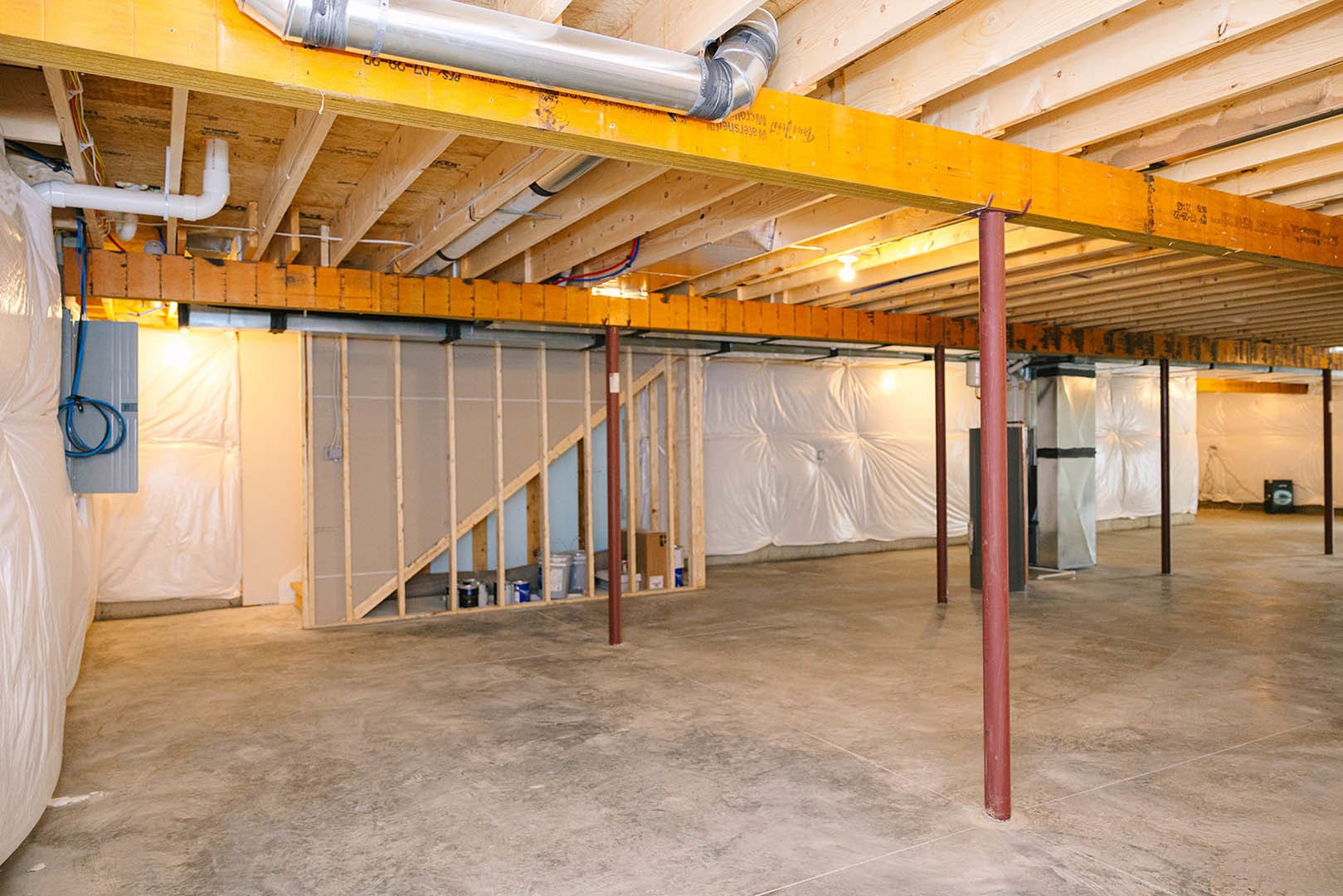 Exposed wooden and steel beams across ceiling, concrete floor, white insulated wall, visible metal pipes and red support pole in unfinished basement room