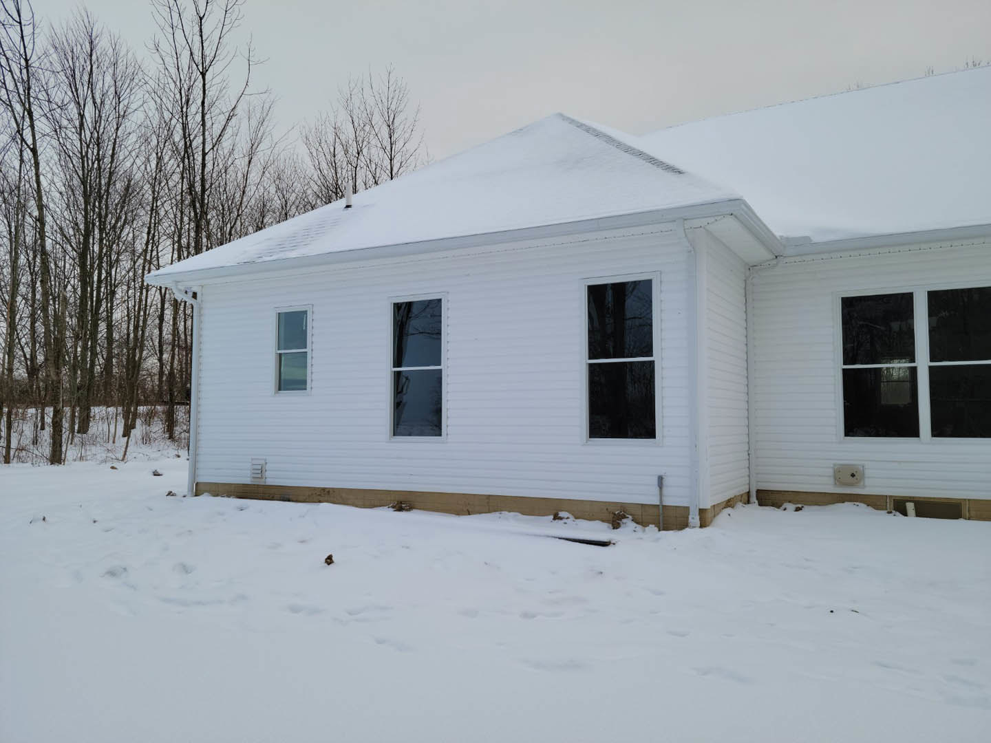 Two-story home with white-framed windows, light siding, and snow covering the roof and ground; bare trees and a visible drainage pipe in the yard.