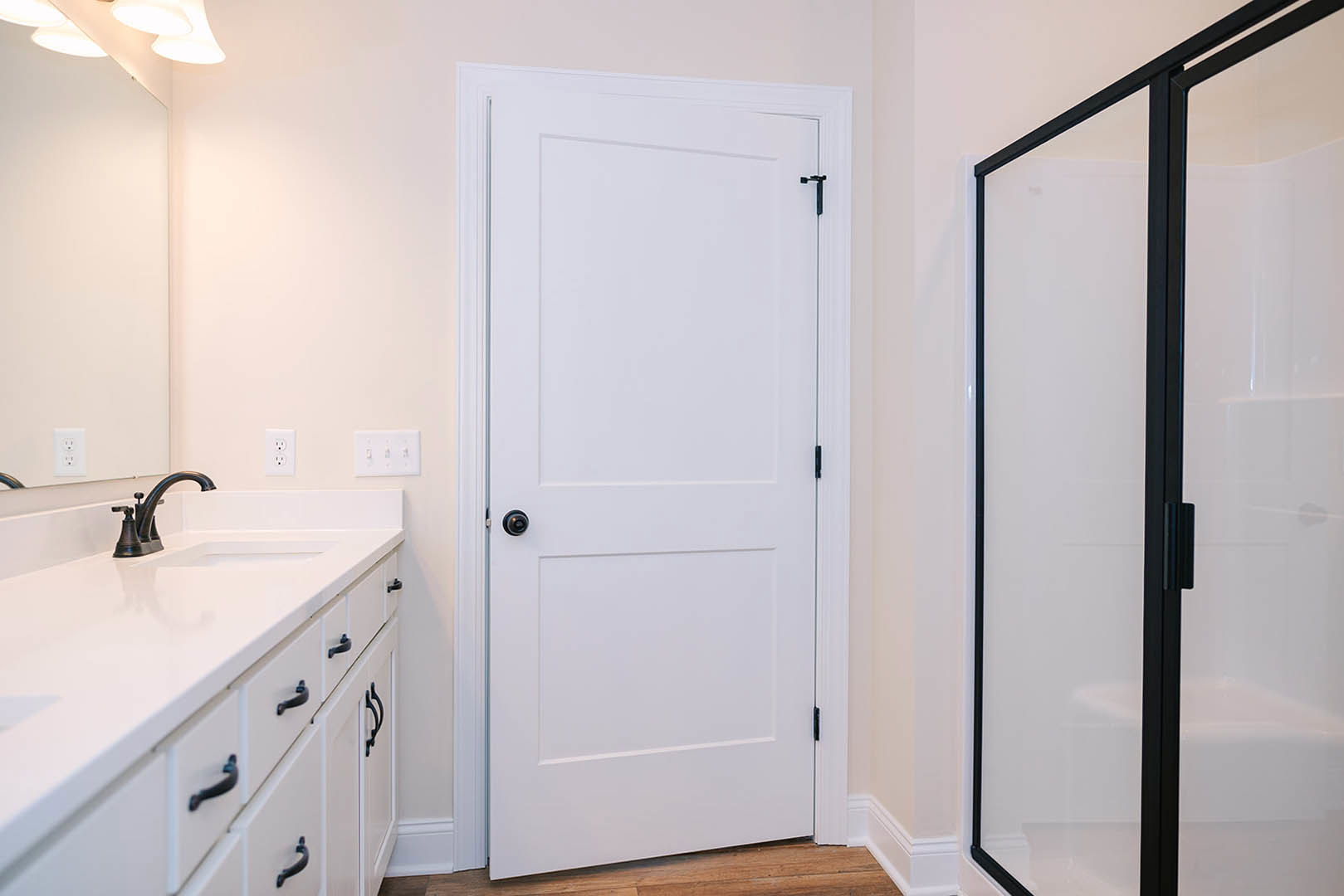 White bathroom door with black knob, white tile walls, black faucet, white countertop, and glass shower door with black handle