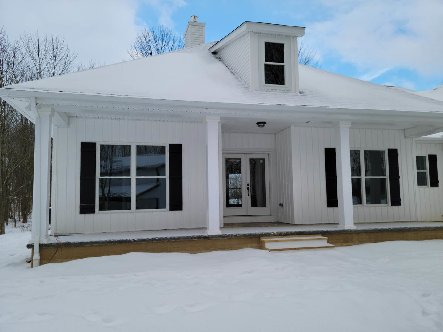 Two-story home with white siding and large windows, snow covering the ground and porch, white-framed windows, wooden bench partially buried in snow, winter landscape.