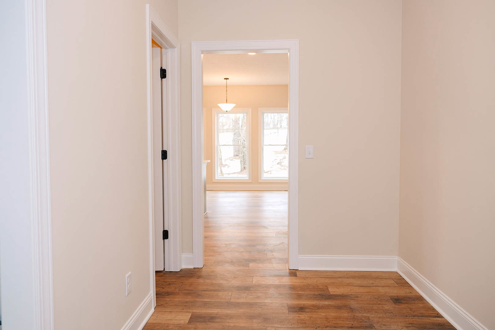 Hallway with warm wood flooring, white-framed window showing snowy landscape, ceiling light fixture, and a door at the end