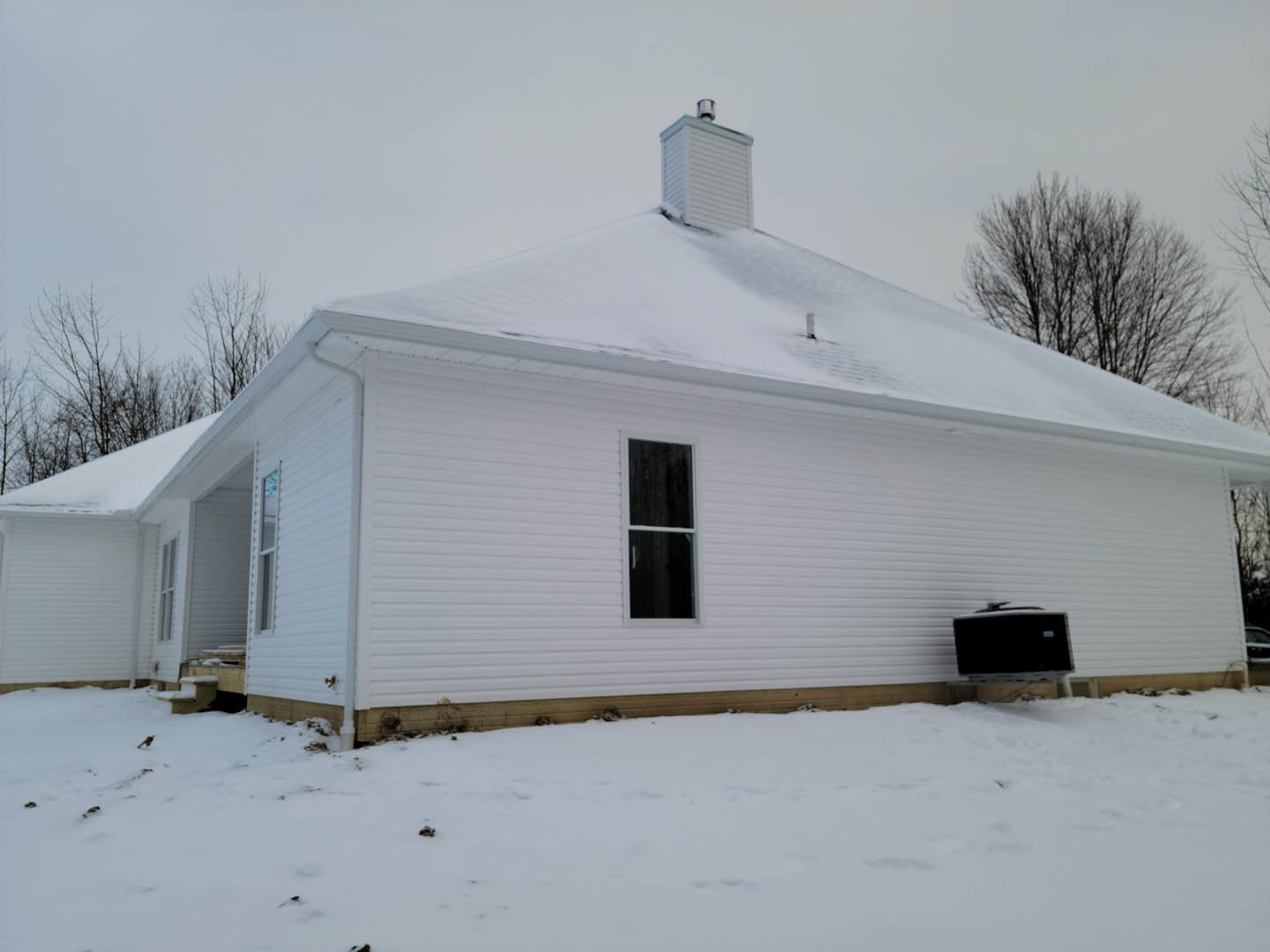White cottage-style home with snow-covered roof and ground, white-framed windows, bare trees, and clear winter sky.