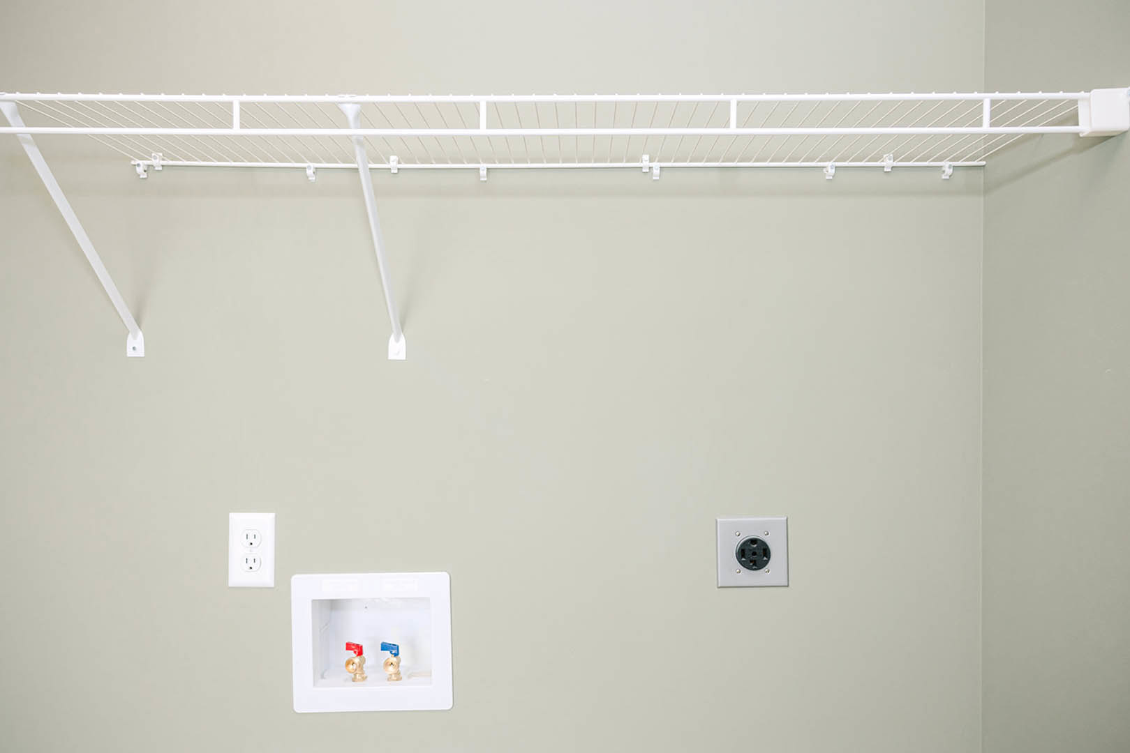 White floating shelf mounted above a smooth painted wall, with a recessed power outlet and a white utility box featuring visible valves below; nearby, a perforated white rack is