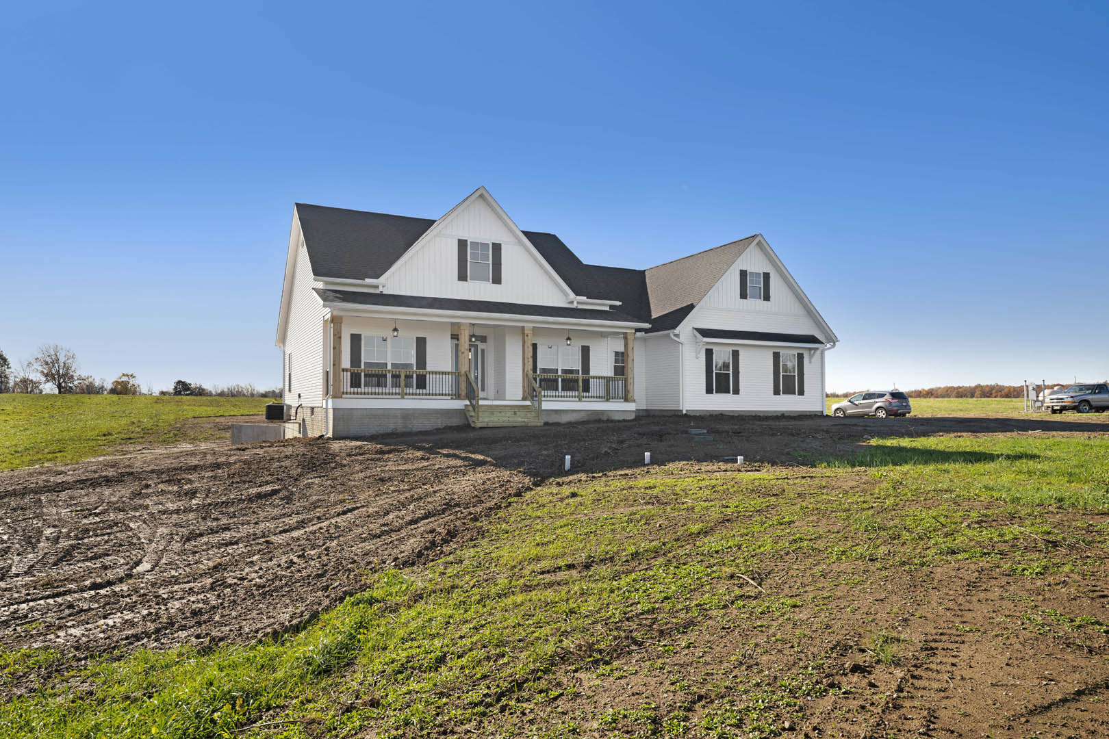 Large white farmhouse with black roof, covered porch with railing, windows, and a dirt field with patches of grass in the foreground; silver truck parked beside the house.