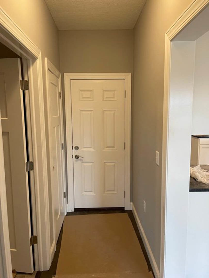 Hallway with white walls, white door featuring a silver handle, patterned rug on light flooring, black countertop against the wall, white plastic bag on the floor, brown paper on a