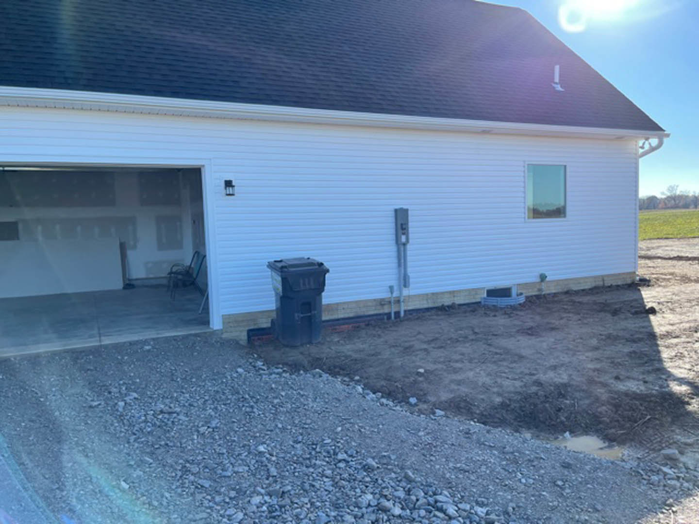 White house with open front door, attached garage featuring a chair and table, gravel driveway, black lidded trash can, pile of rocks beside building, blue sky with scattered