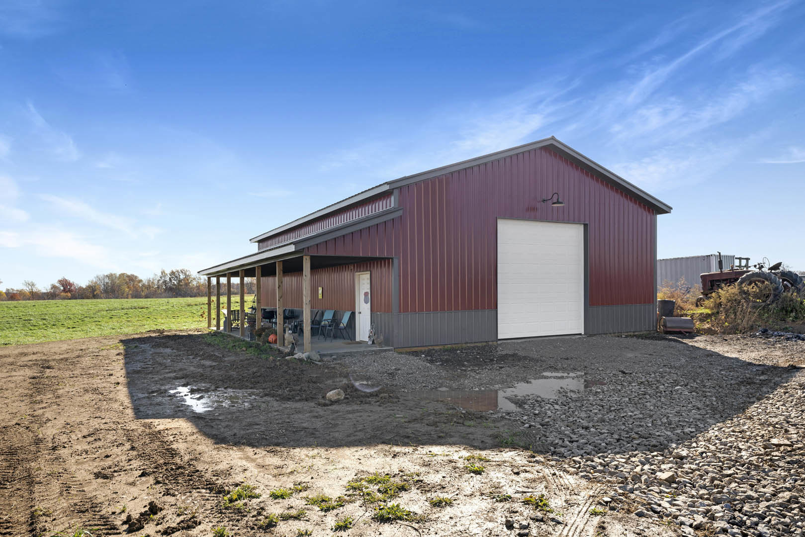 Red barn-style home with covered porch, white garage door with black trim, front door flanked by outdoor chairs, muddy yard with puddle, grassy field, and scattered trees under