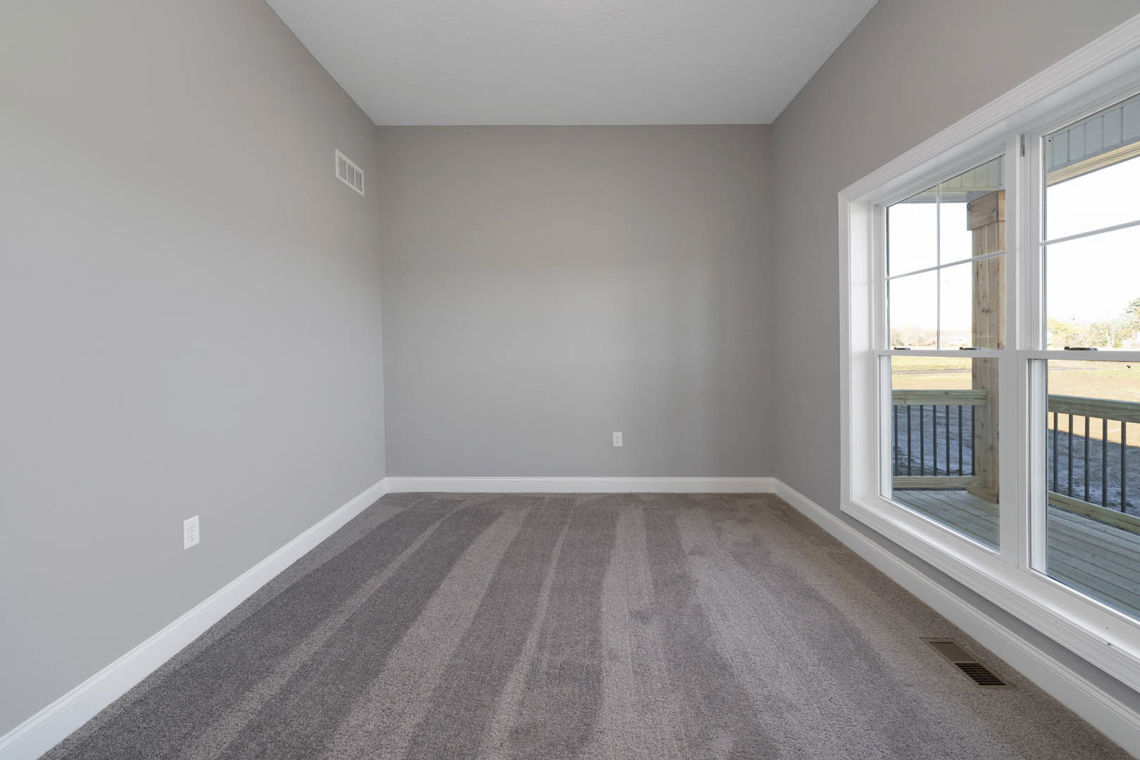 Carpeted room with white walls, black baseboard trim, large window overlooking yard, white floor vent near wall