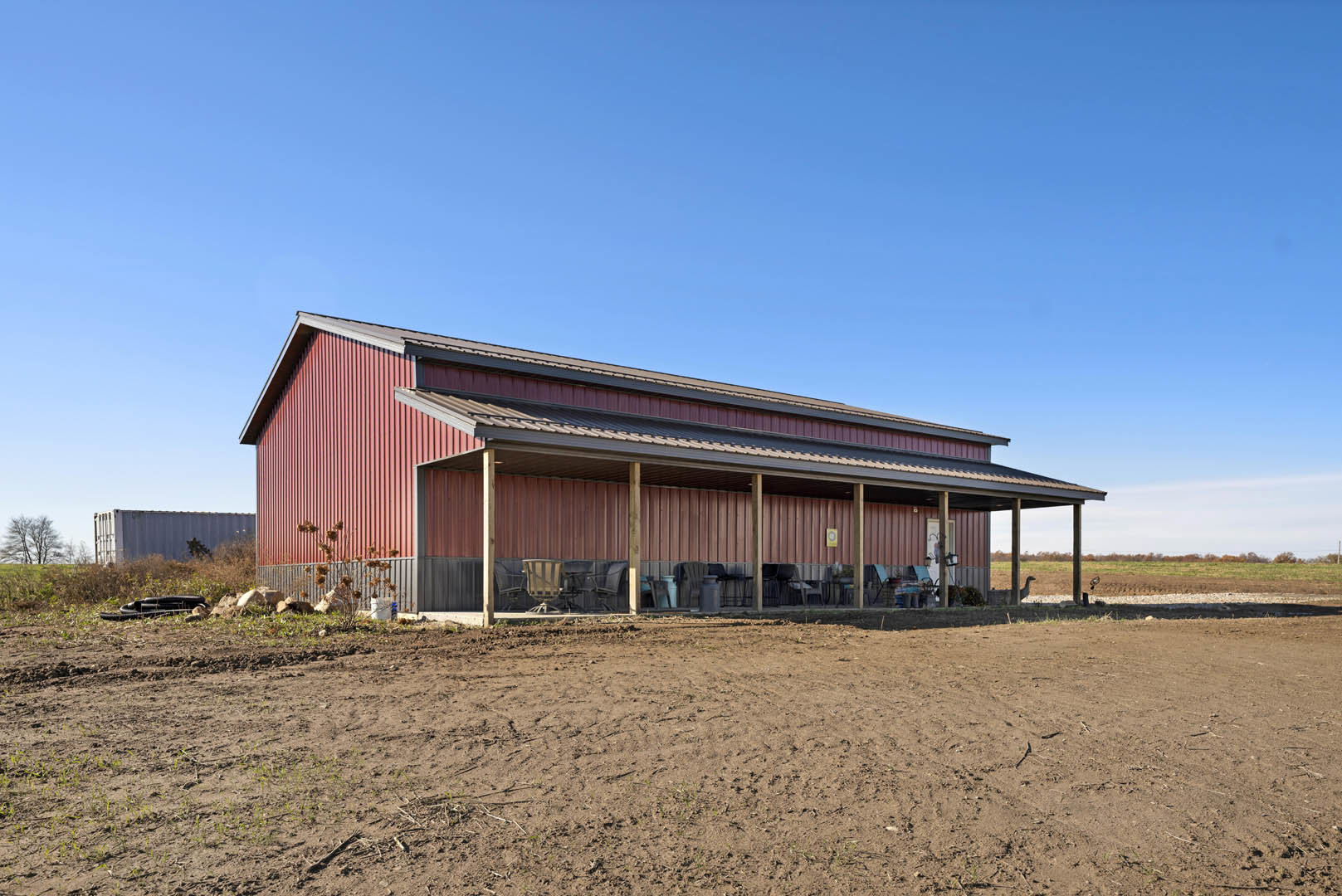 Red-painted house with covered porch, white trim, metal roof, dirt yard with scattered chairs, wooden fence, and surrounding trees.
