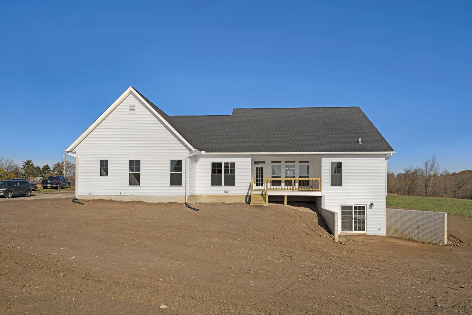 White siding house with a wooden ramp leading to the front porch, elevated deck, brick retaining wall, dirt and grass landscaping, blue sky overhead, car parked roadside.