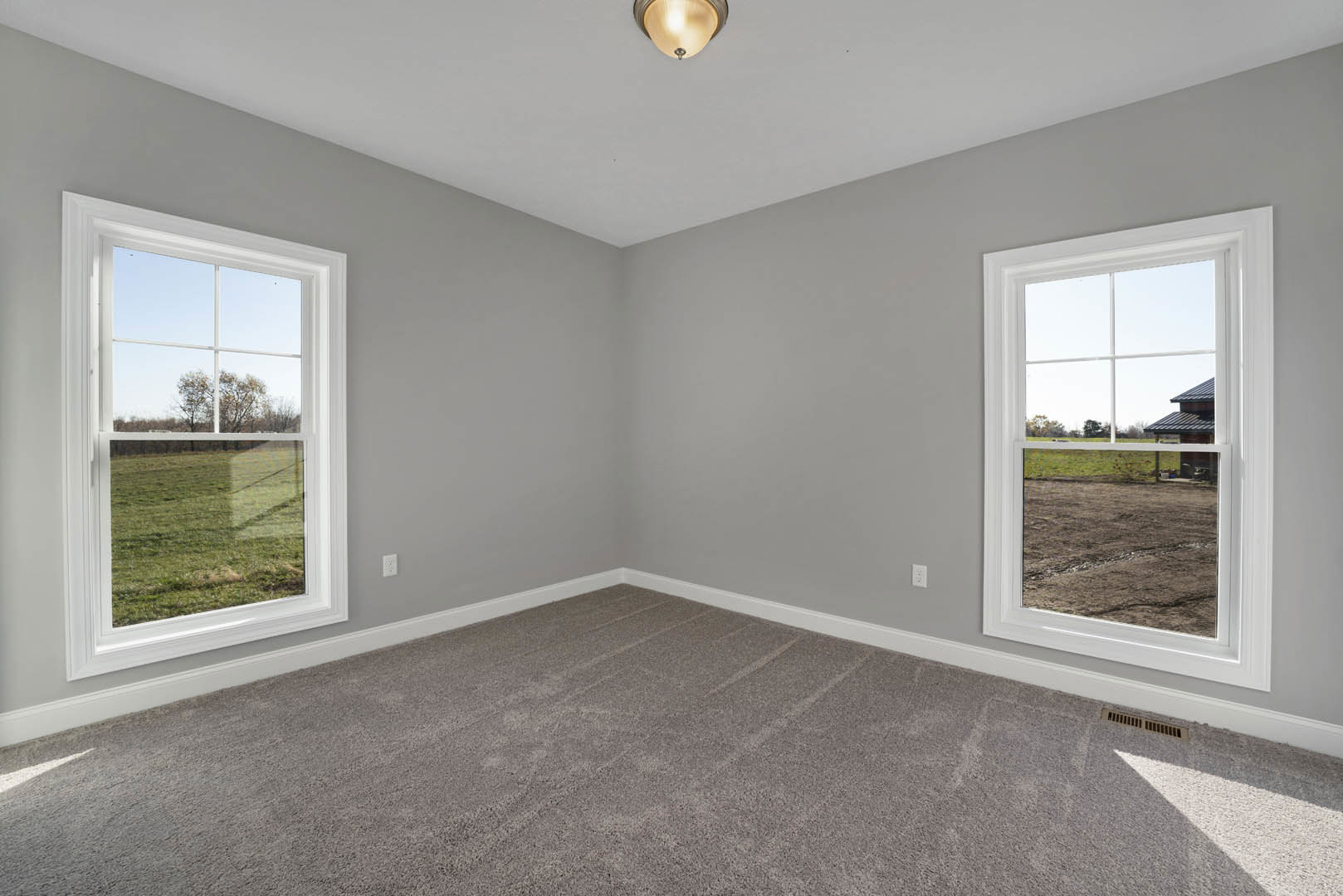 Carpeted room with large windows, white walls, ceiling light fixture, and view of open field and farm buildings through the windows