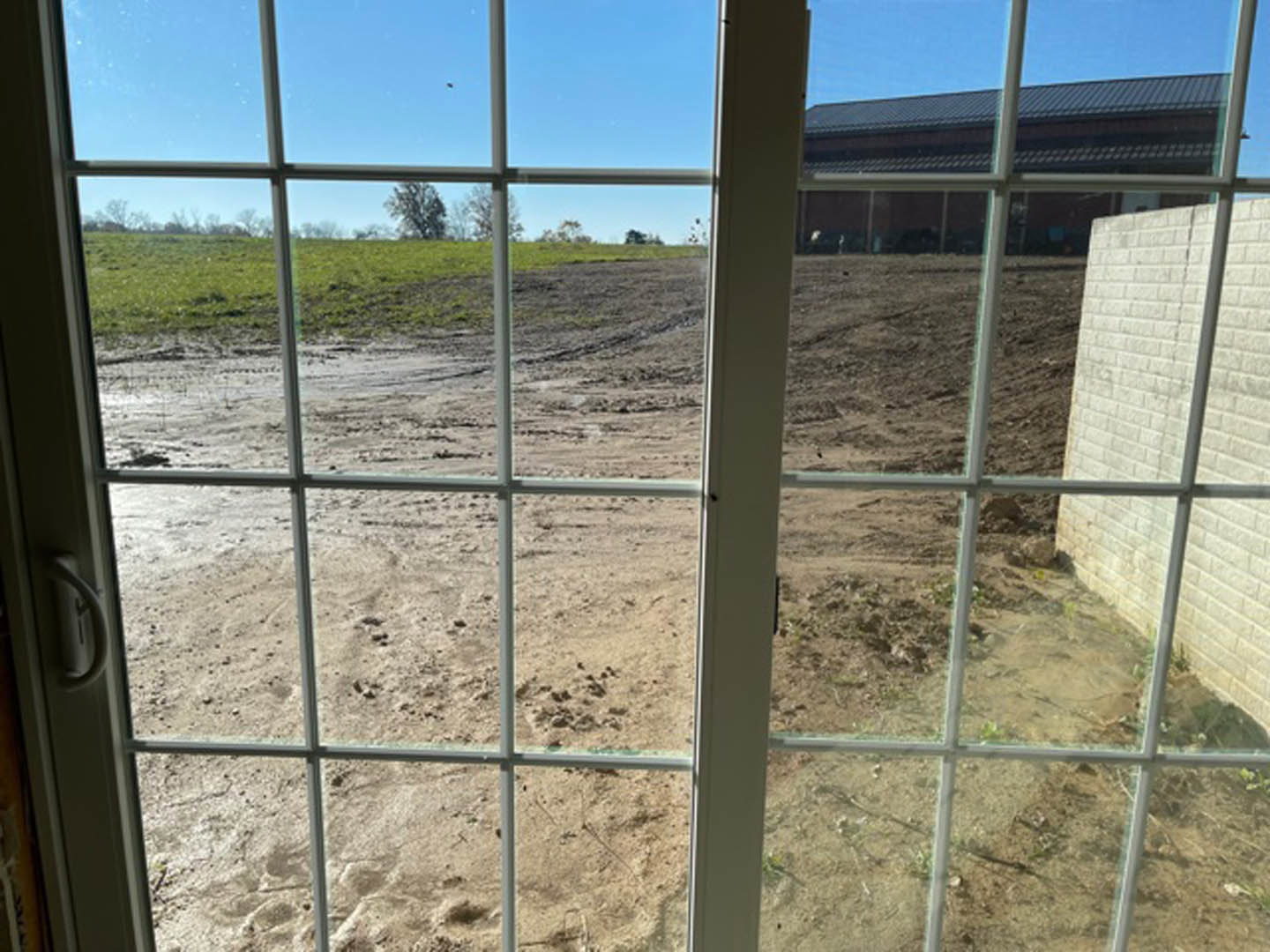 White brick window frame overlooking a dirt field with patches of grass, wire fencing, and distant trees under a clear blue sky.