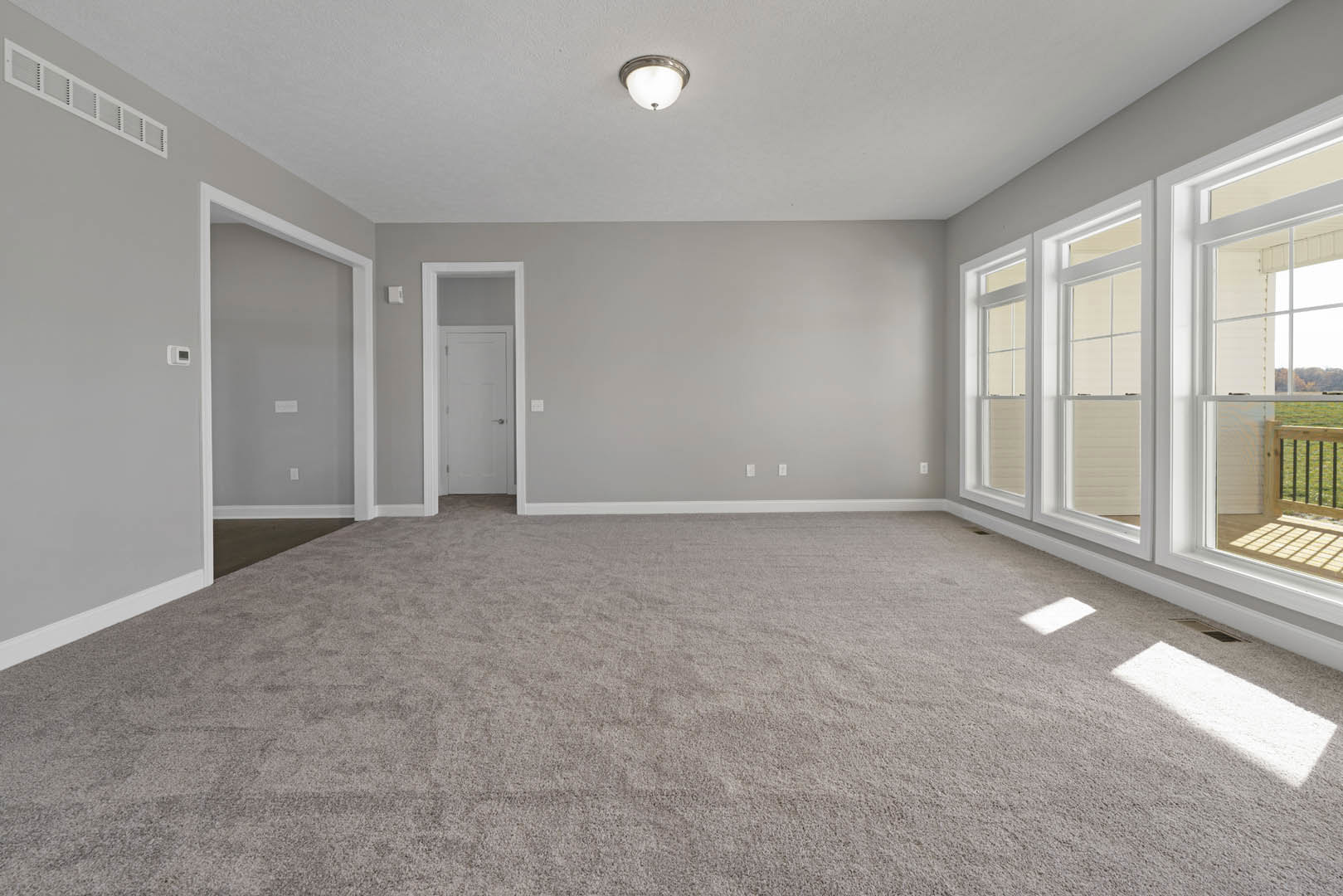 Bright carpeted room with large windows, white walls, ceiling light fixture, white door with silver handle, sunlight streaming onto pale carpet, view of deck through windows