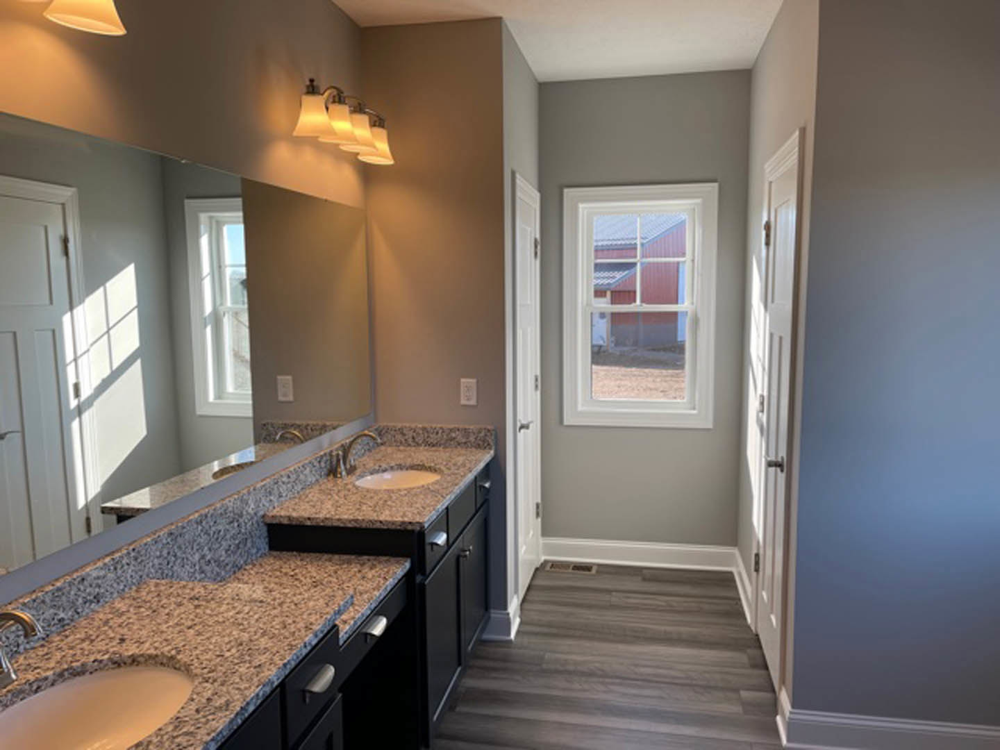 Granite bathroom countertop with undermount sink, large framed mirror above, row of wall-mounted lights, wood flooring, window revealing barn outside, white ceramic bowl on