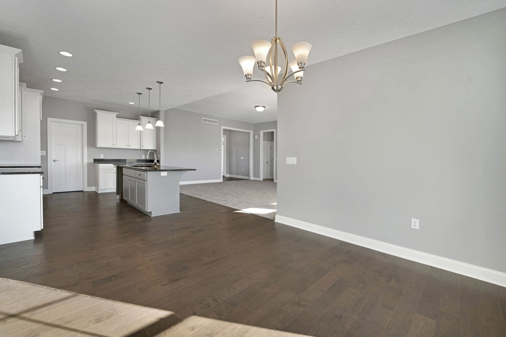 Open kitchen and dining area with hardwood flooring, white cabinetry, kitchen island featuring a sink, modern chandelier overhead, white door with silver handle and nearby light