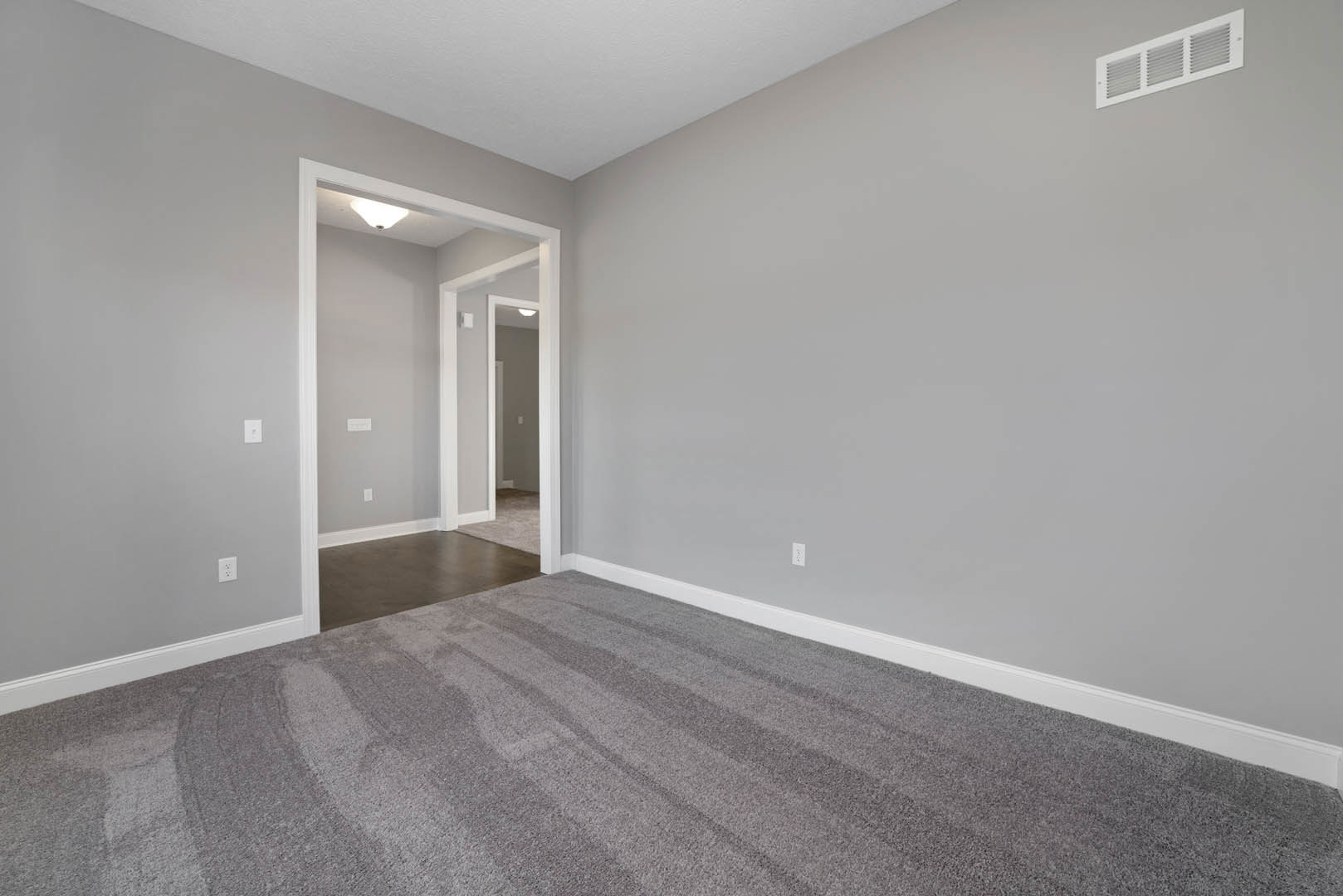 Open white door leading to carpeted room with grey flooring, white walls, hallway with white trim, wall vent, and plaster ceiling.