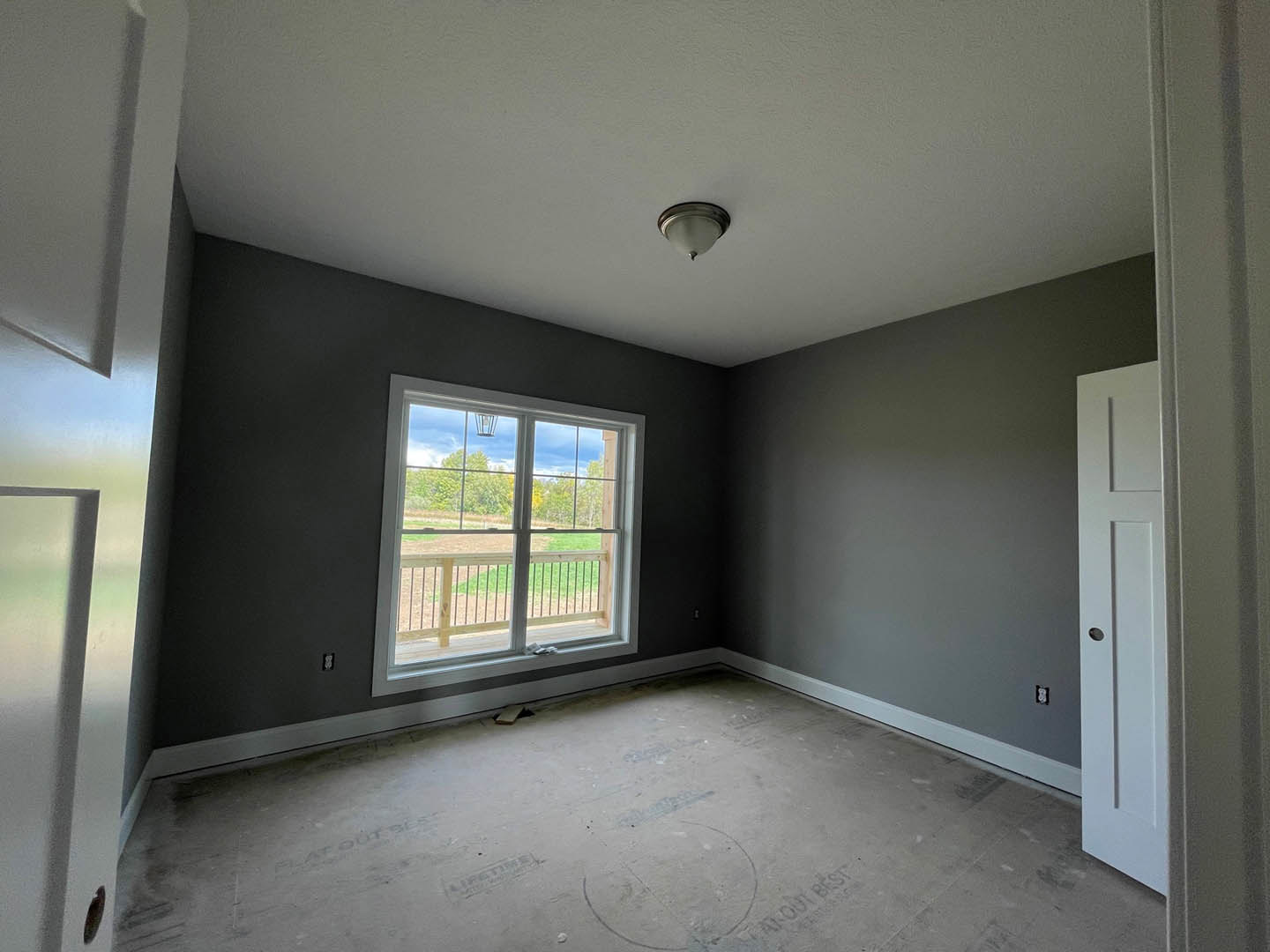 Modern interior room with plaster walls, large window overlooking fenced grassy yard, close-up of white door, concrete floor featuring circular pattern, ceiling-mounted light
