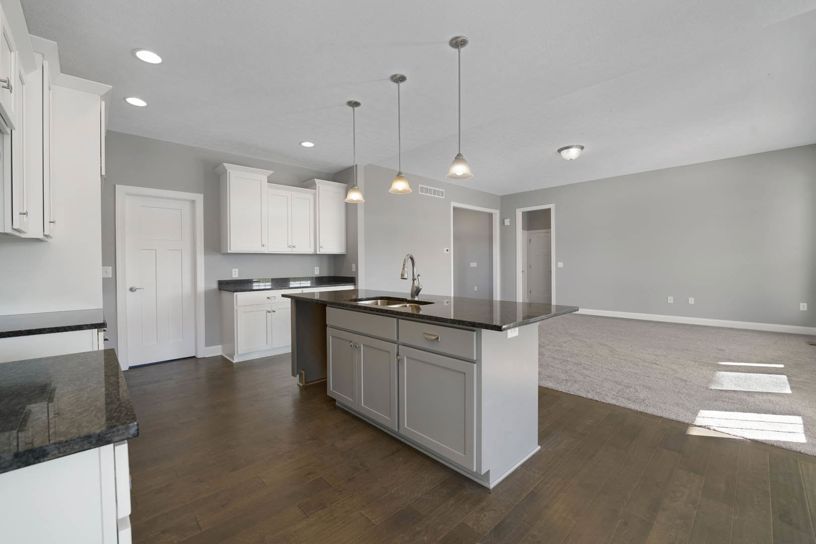 Open kitchen and living room featuring white cabinetry, white door with silver handle, kitchen island with built-in sink, dark wood flooring, and light countertops.