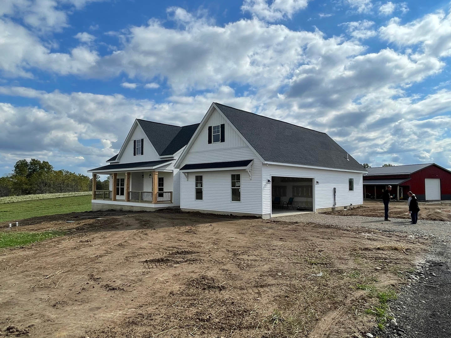 Two-story house with attached garage, light siding, gable roof, and large windows, set on a dirt lot with grass and trees under a blue sky with clouds