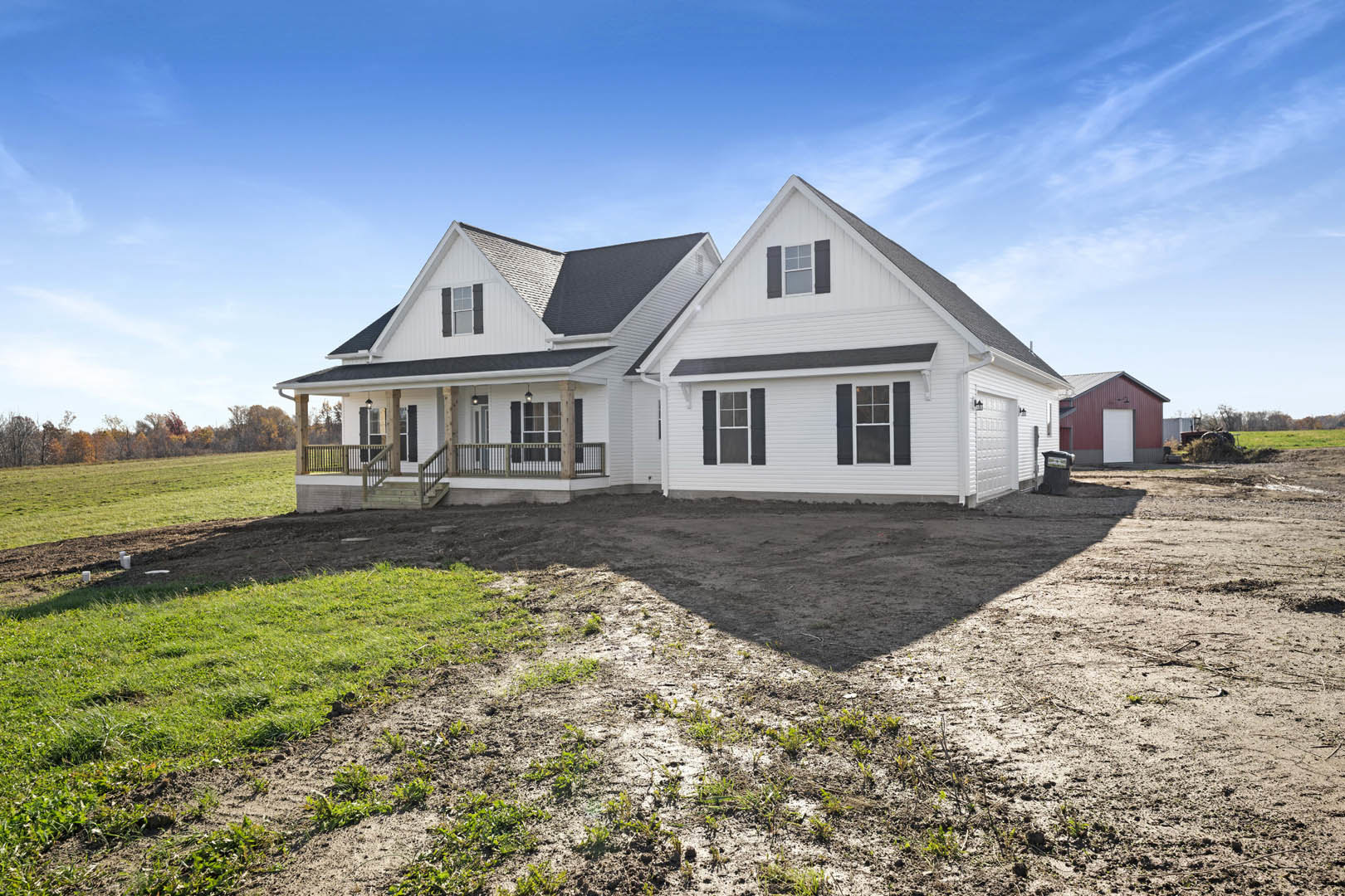 White house with black shutters and black roof, surrounded by dirt and patches of grass, red barn with white door in background, wooden deck with railing attached to the house