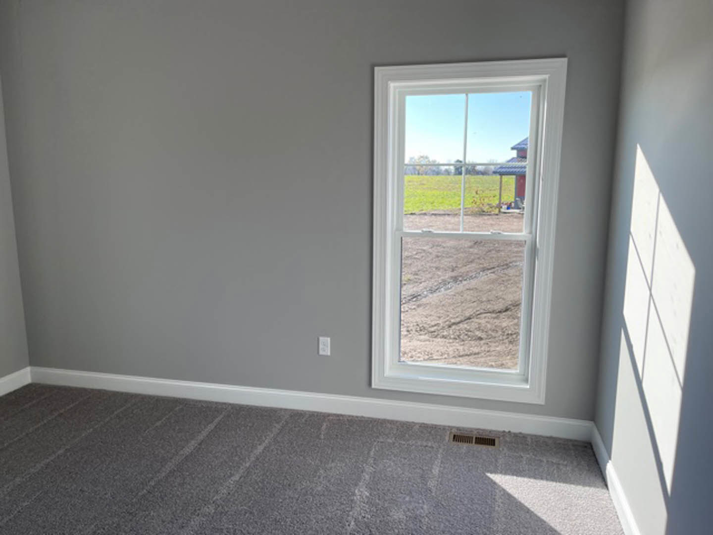 Sunlit room with grey carpet flooring, large window framed in white trim, plaster walls, and view of grassy field and distant house outside