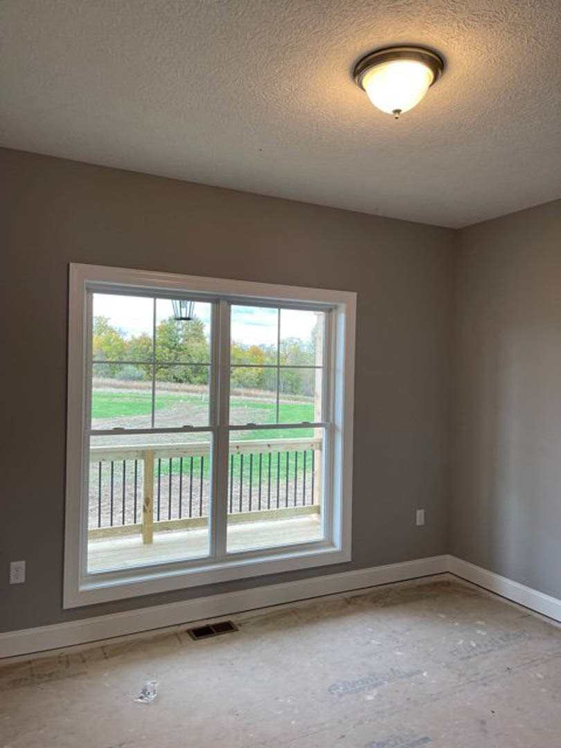 Bright room with white plaster walls, large window overlooking fenced grassy yard, wood floor with central vent, ceiling light fixture, balcony railing visible through glass.