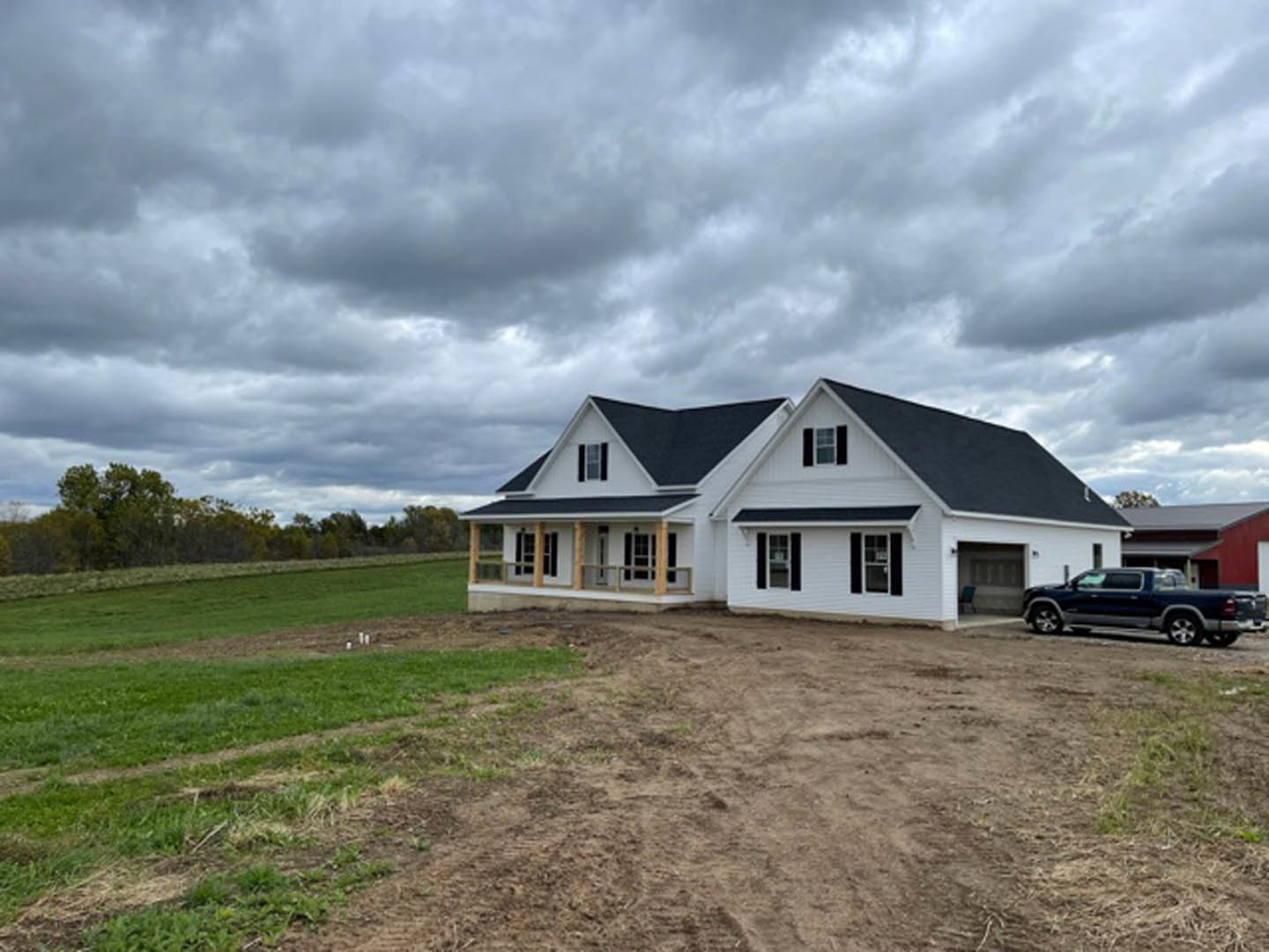 Two-story custom home with gray siding and white trim, parked pickup truck in gravel driveway, unfinished landscaping, cloudy sky overhead