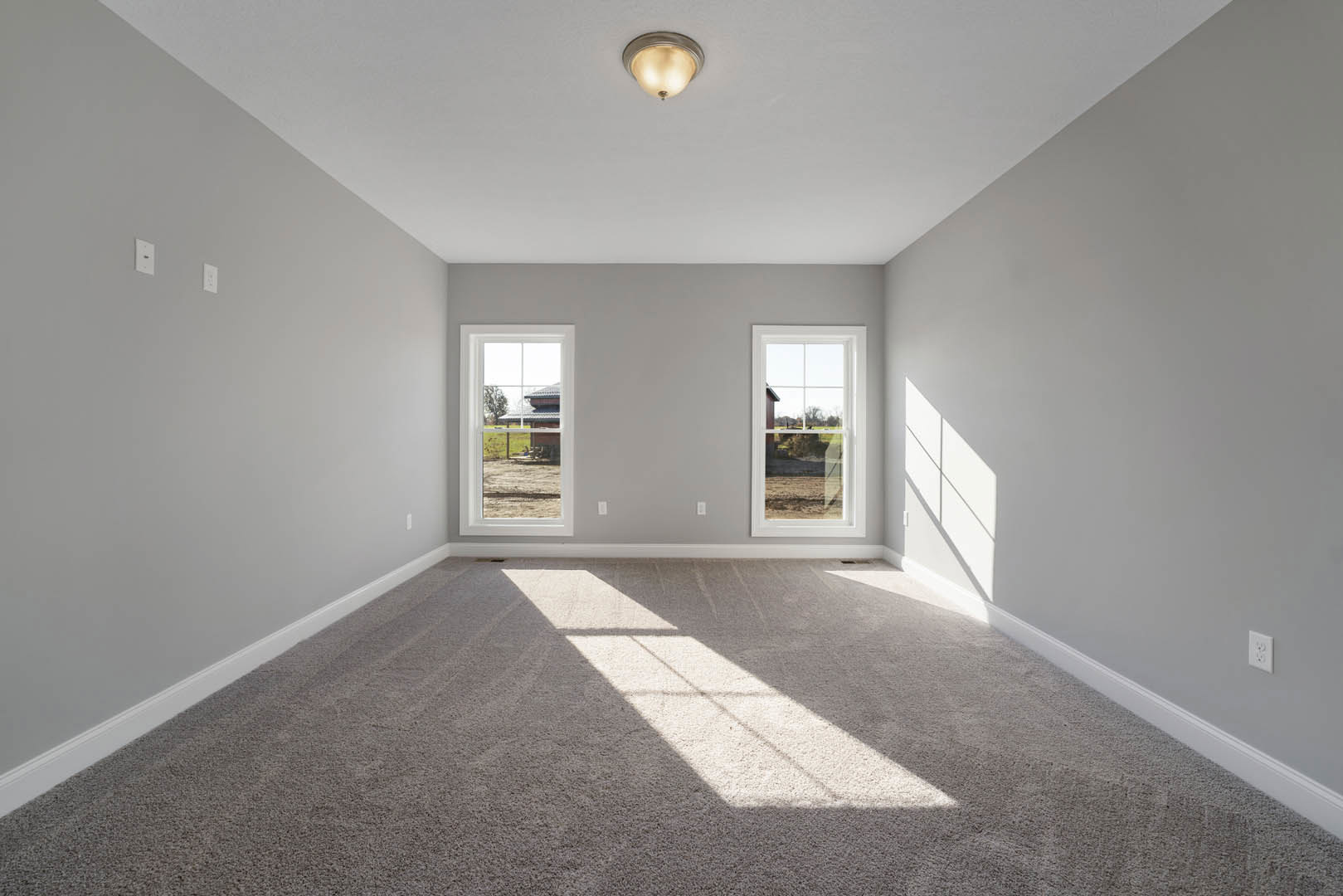 Carpeted room with two large windows, white walls, ceiling light fixture, and views of a farm and neighboring house through the windows