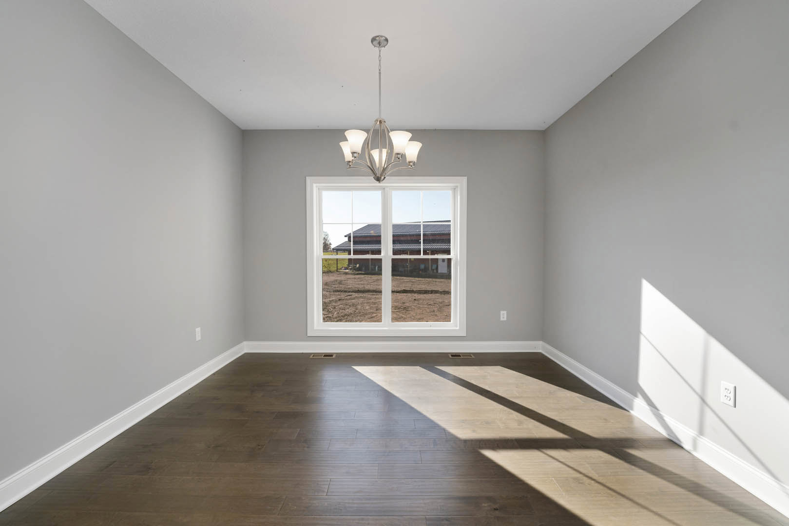 Chandelier hanging from ceiling in a room with hardwood floors, white walls, and large window overlooking farmland and a distant building