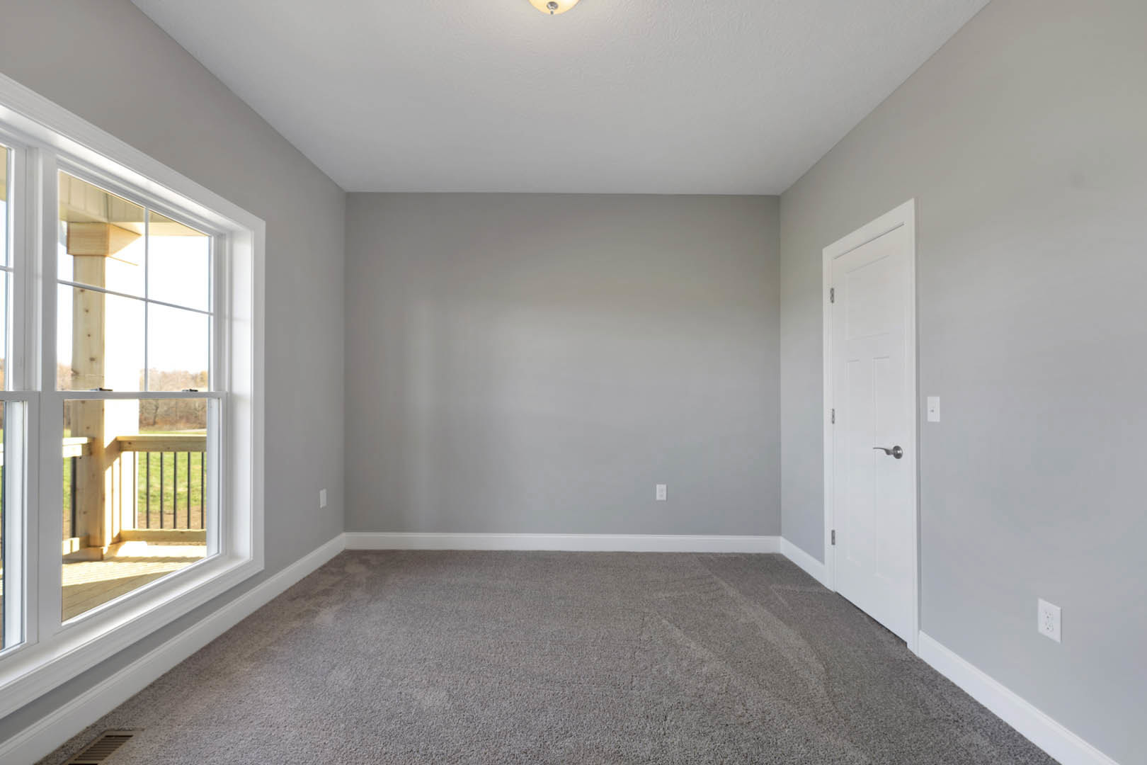 Carpeted room with white walls, open white door with silver handle, large window overlooking grassy field, simple baseboard molding