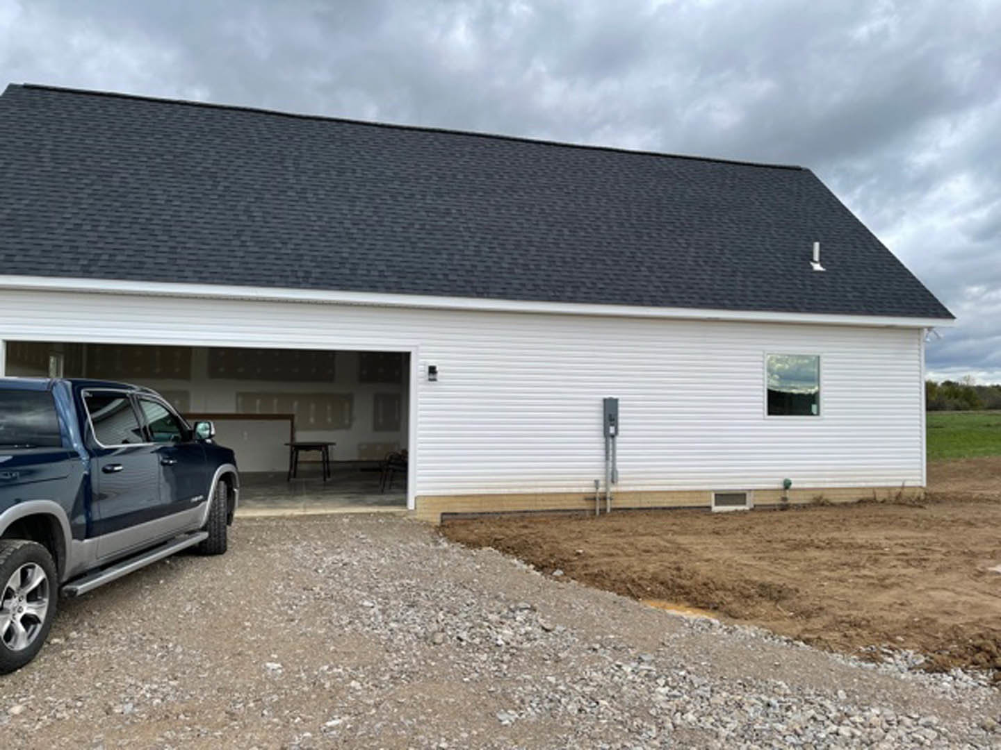 White-framed window and gray roof above a garage with a parked truck, tire visible, bordered by a dirt patch and cloudy sky