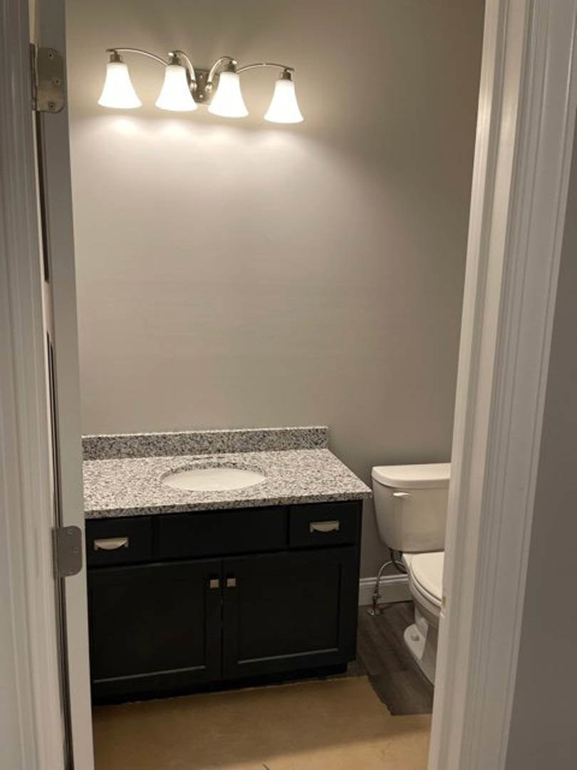 Marble countertop with undermount sink, black cabinet, chrome faucet, white toilet, and dual light fixture above, set against light tile walls