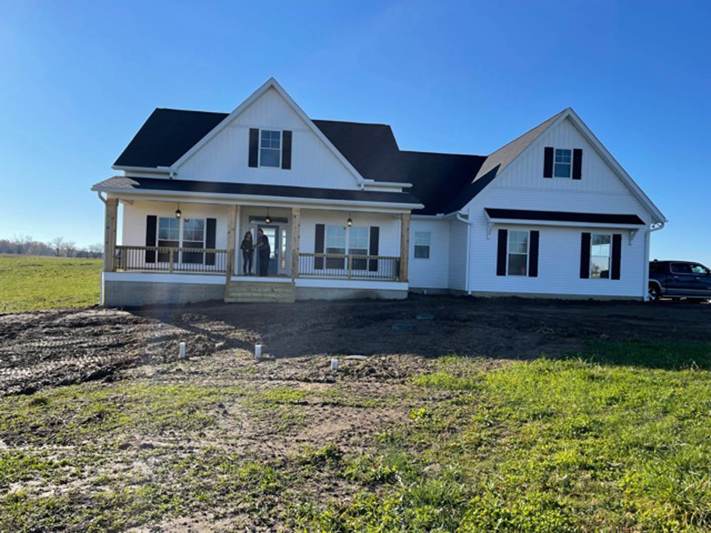 Two-story house with white siding and dark roof, wide front porch, large green lawn, dirt patch near entry, square-paned windows, man and woman shaking hands on porch, parked car
