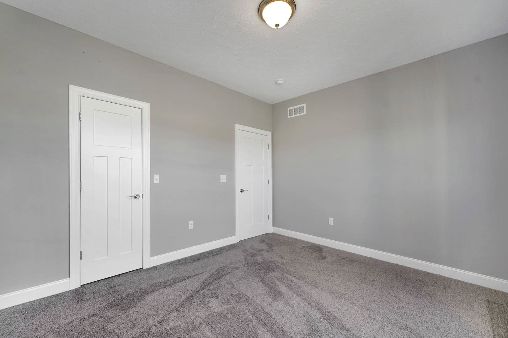 Carpeted room with two white doors, silver handles, ceiling light fixture, wall vent, and white baseboards.