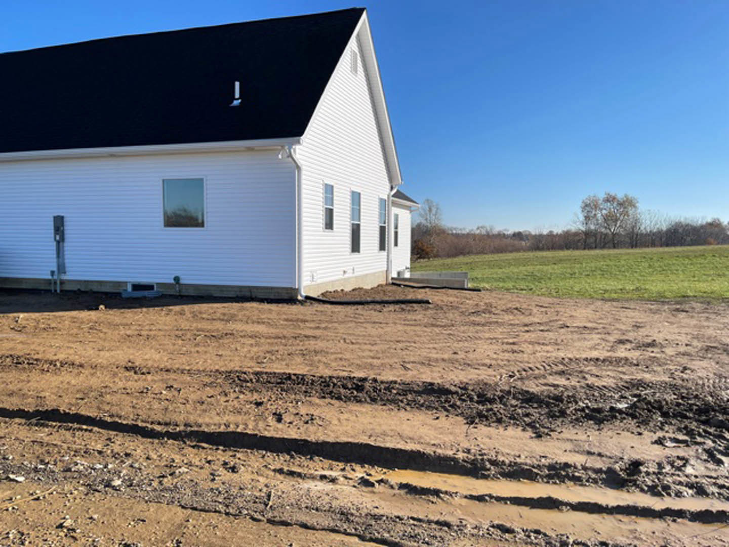 White house with black roof and multiple windows, surrounded by dirt patch and grassy field, trees and blue sky in background, black rectangular sign with white letters near front