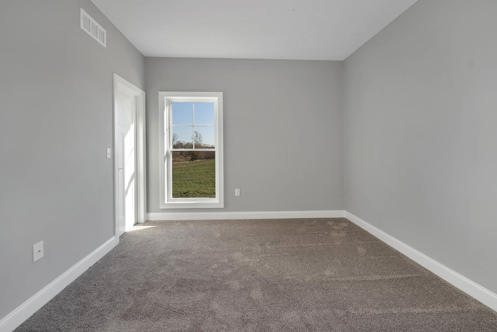 Carpeted room with white walls, large window overlooking grassy field and tree, wall outlet with black button, and wall vent