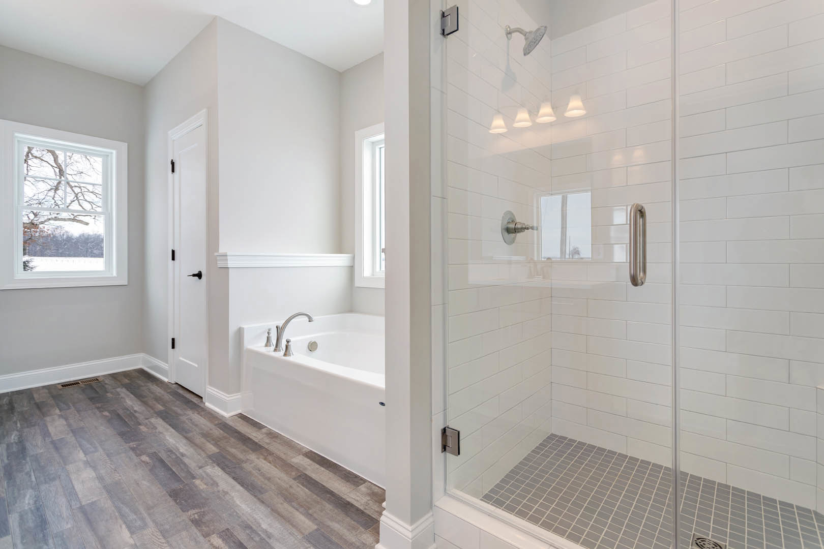 Bathroom with glass shower enclosure, white tile floor, freestanding bathtub, wood flooring with white trim, large window overlooking snowy field, chrome fixtures, and central