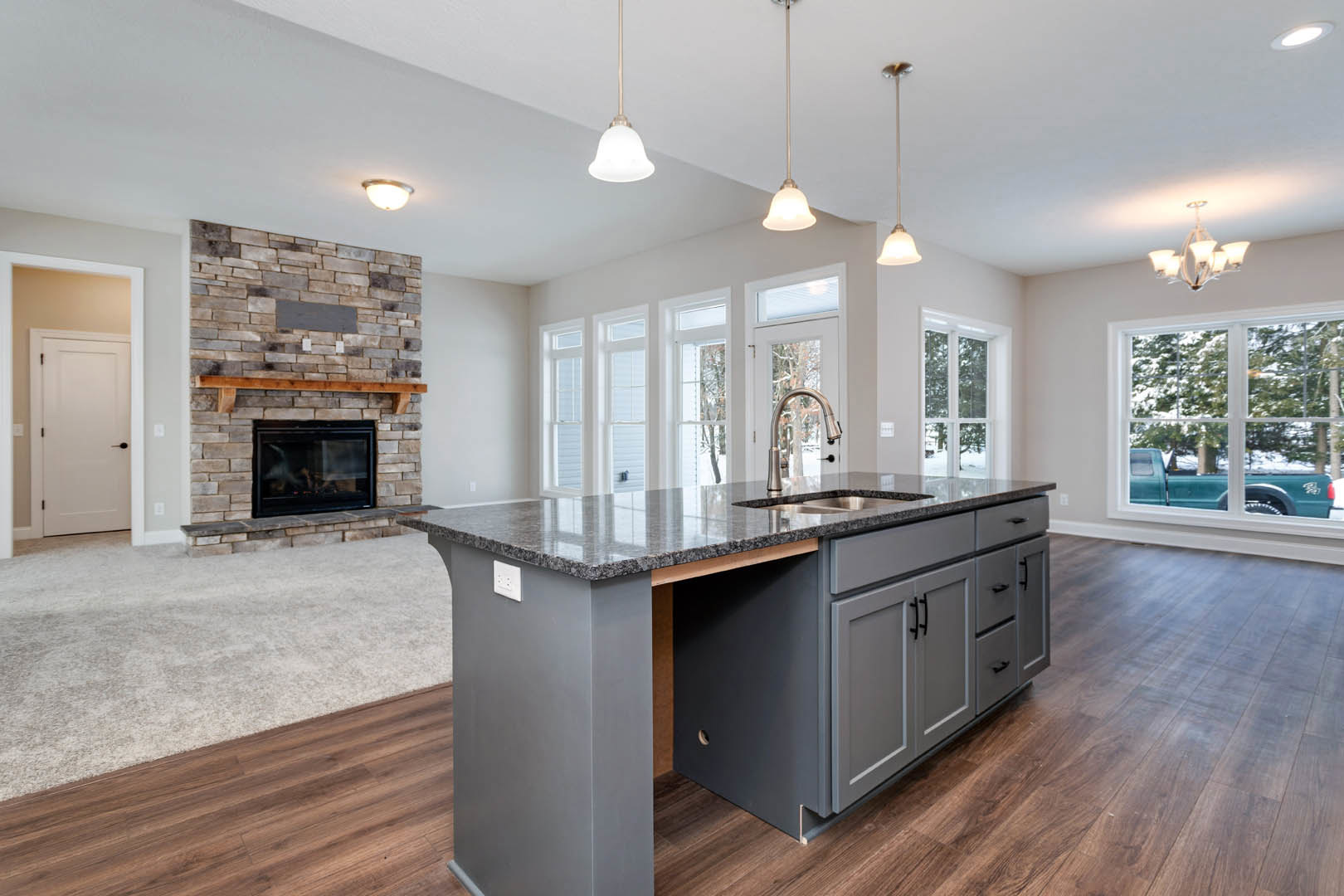 Open-concept kitchen and living room featuring a stone fireplace with wood mantel, white cabinetry, quartz countertops, stainless steel sink, white door with black handle, and
