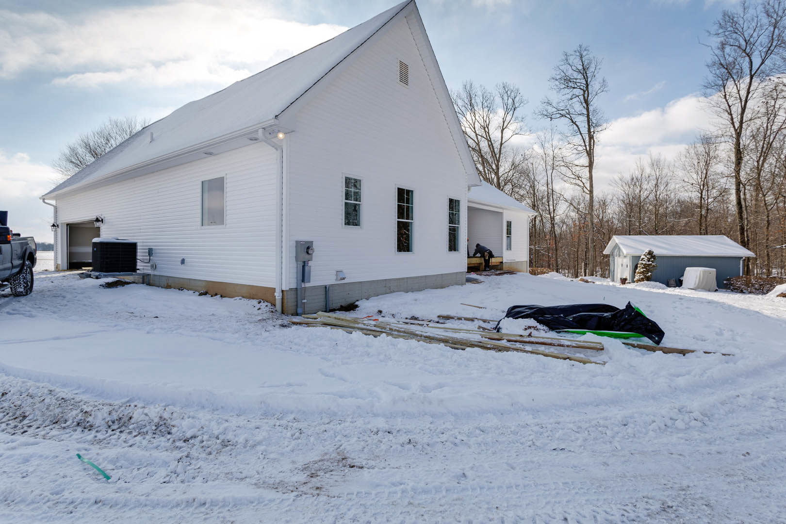 White two-story house with snow covering the lawn, a snow-covered car parked in front, bare tree branches dusted with snow, and a stack of firewood partially buried in snow.