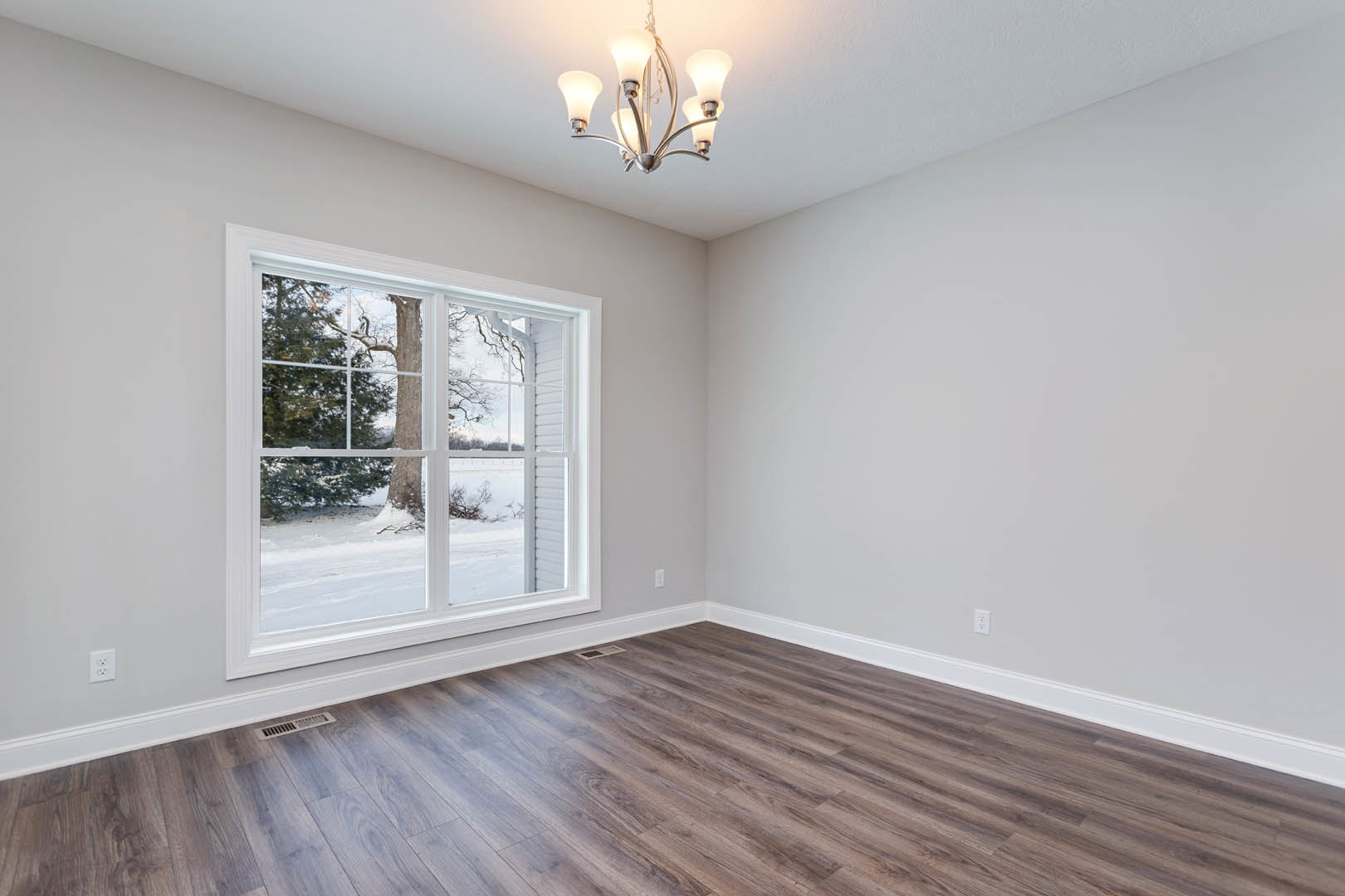Sunlit room with wide window overlooking snowy landscape, wood flooring, white walls with crown molding, and modern five-light ceiling fixture