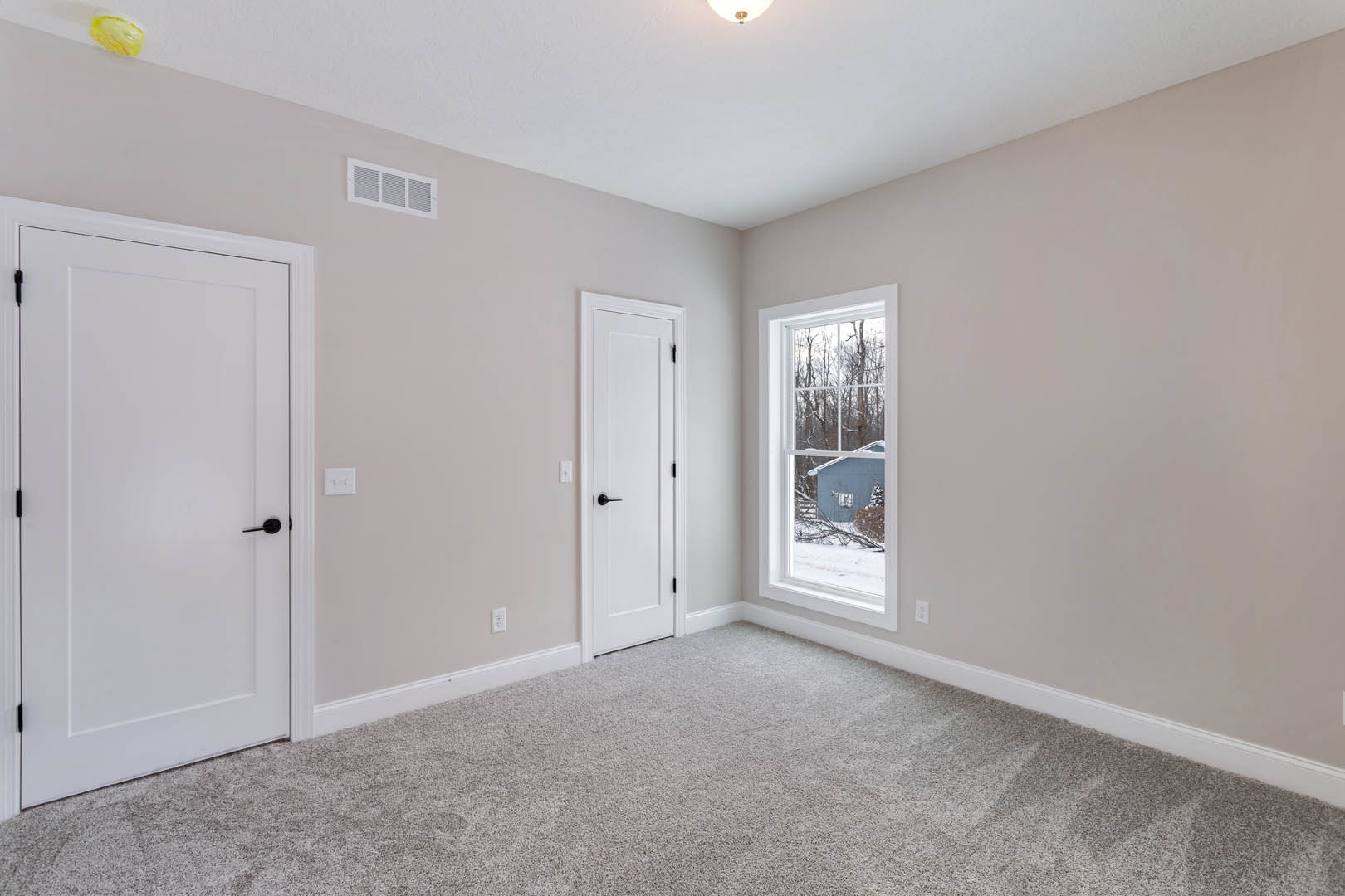 White paneled doors with black handles, carpeted walls, white baseboards, a yellow item on a white shelf, window overlooking neighboring house, light carpeted floor.