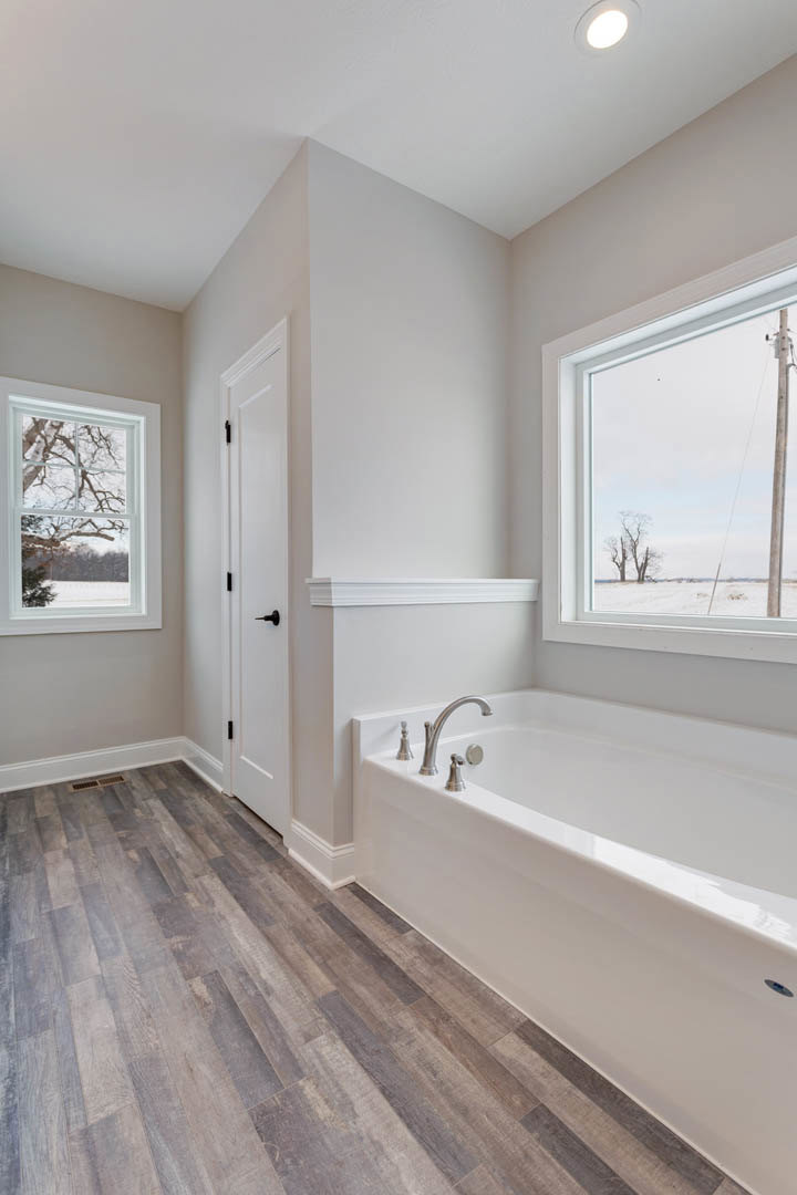Freestanding bathtub with chrome faucets on wood flooring, two large windows with views of trees, white door with black handle, light plaster walls, decorative molding
