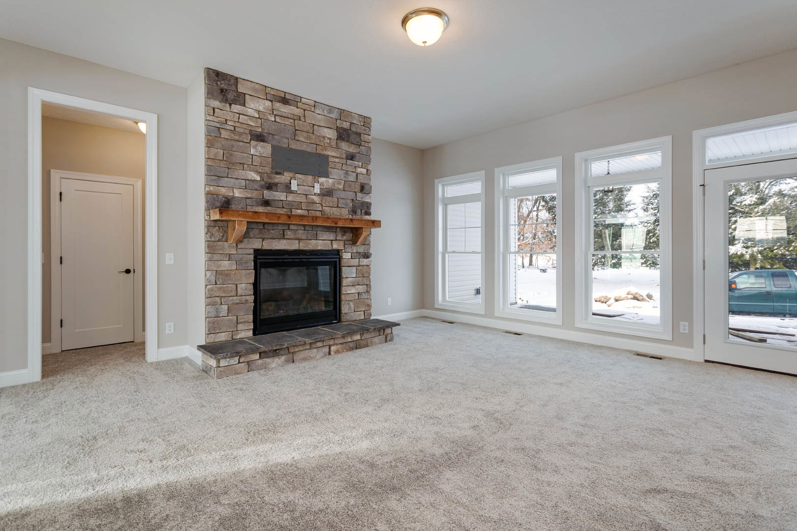 Living room with beige carpet, white walls, glass-door fireplace with wood mantel, ceiling light fixture, and white door with black handle