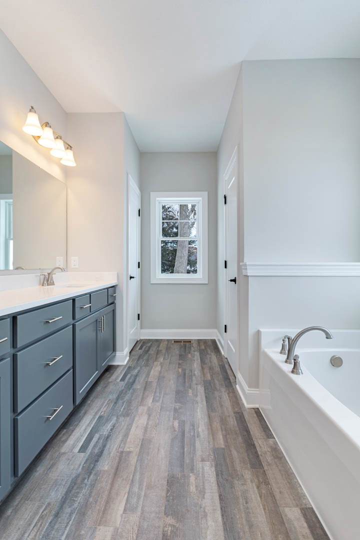 Freestanding bathtub and rectangular sink on stone countertop, white tile walls, chrome faucet, large window with tree trunk view, wood floor, recessed lighting