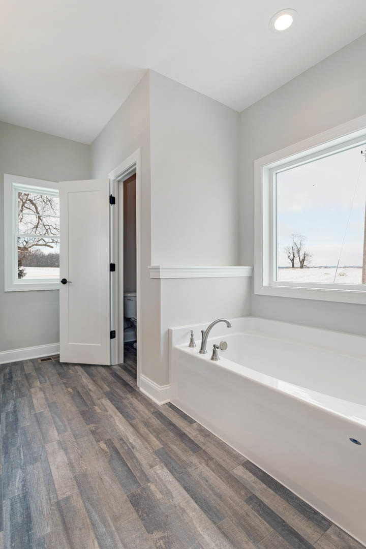 Bathroom with freestanding white bathtub, wood flooring, white door with black handle, ceiling light, window showing tree outside