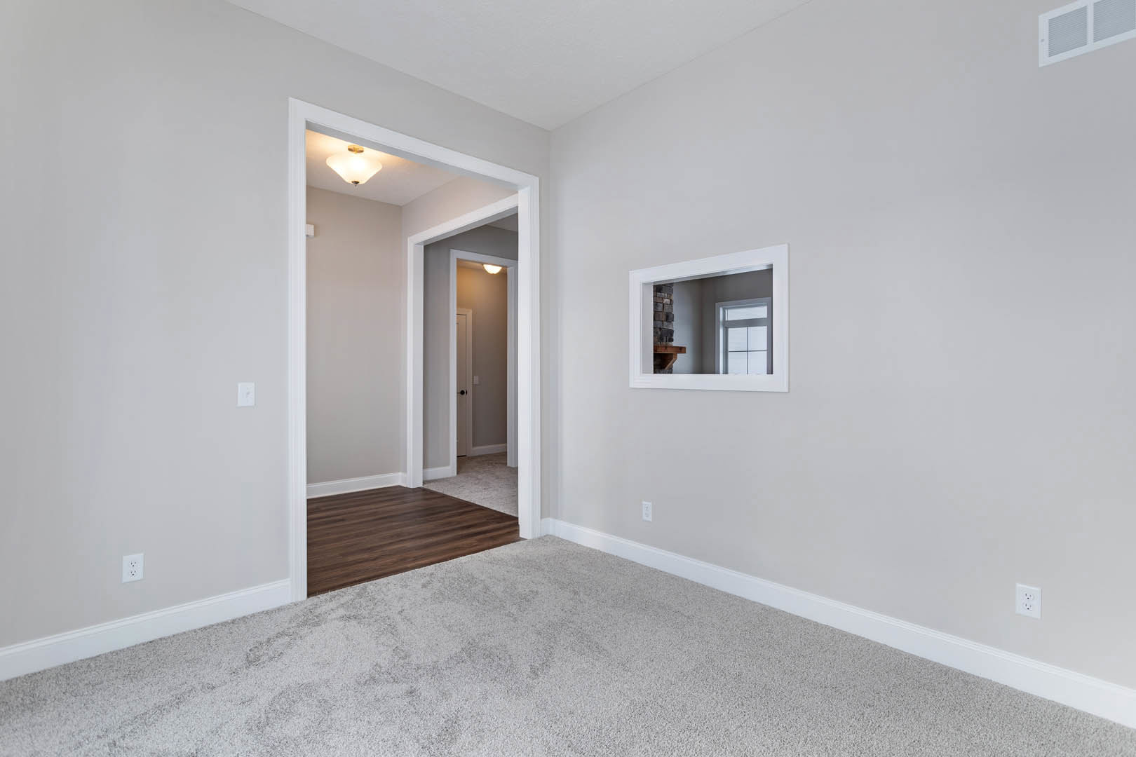 Open doorway leading to a room with wood flooring, white carpet, wall mirror reflecting a fireplace, window, and ceiling light fixture