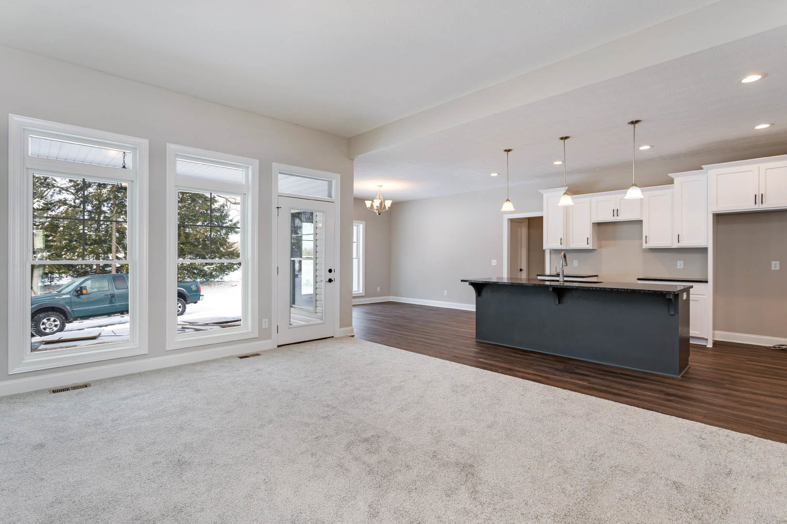 Open kitchen with black countertop, light cabinetry, large window overlooking snowy driveway with green truck, adjacent door, and carpeted area