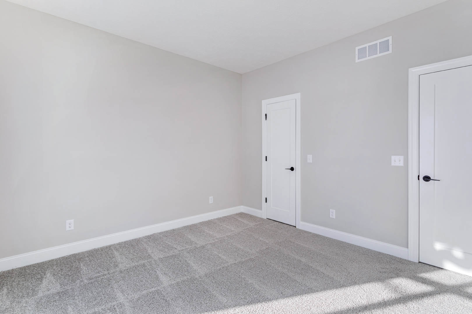 White paneled door with matte black handle, beige carpeted floor featuring subtle white square pattern, wall-mounted vent, smooth white walls, soft natural light filtering in.